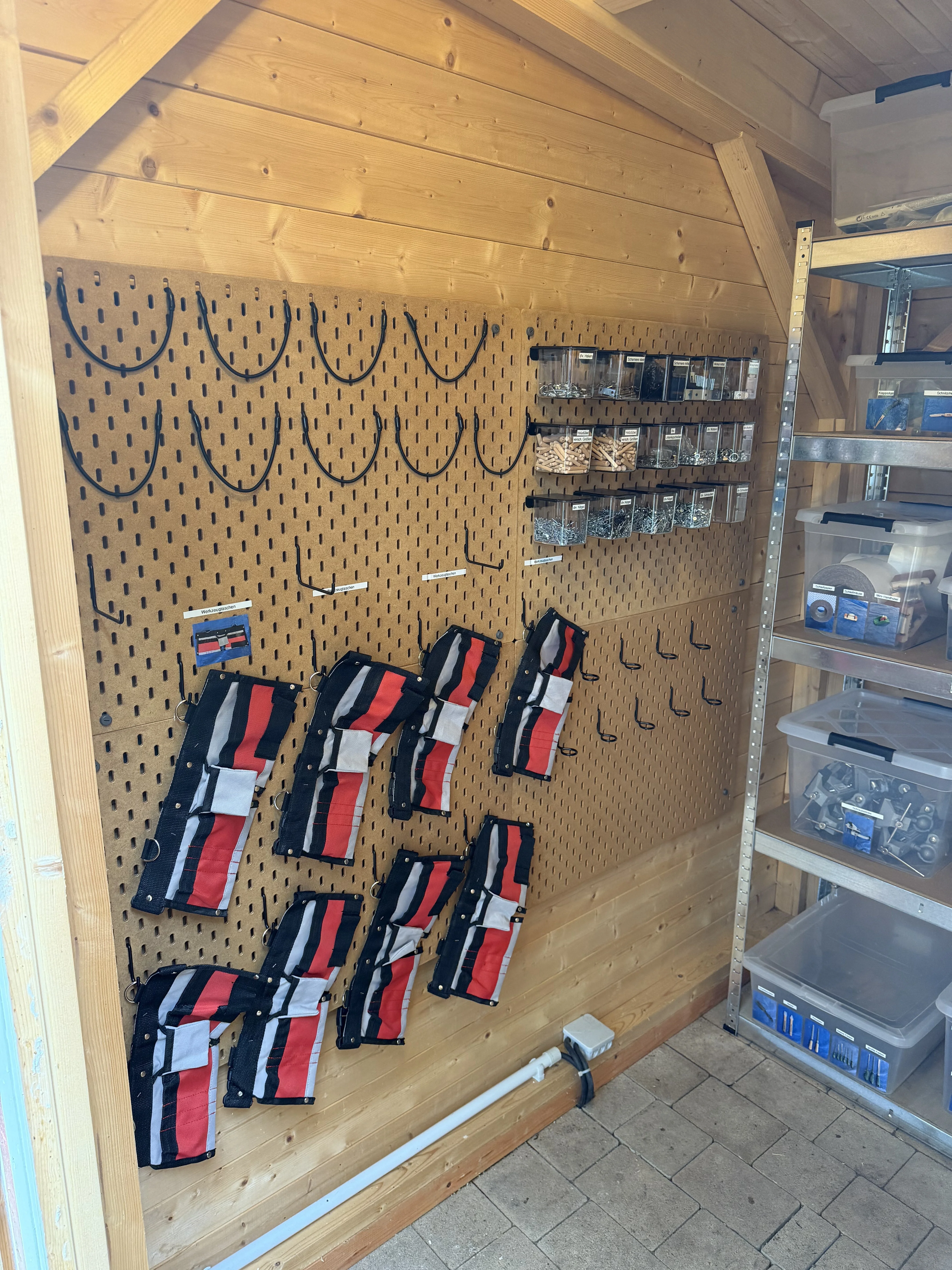 Auf dem Bild ist eine gut organisierte Werkstatt- bzw. Lagerwand zu sehen: Eine große Holz-Pegboardwand mit zahlreichen Haken und Aufhängungen. Mehrere rot-schwarz-weiße Werkzeug- oder Schutztaschen hängen daran. Oben rechts befinden sich transparente Sortierboxen mit Kleinteilen wie Schrauben, Dübeln und Beschlägen. Daneben steht ein Metallregal mit weiteren Kunststoffboxen und Material. Die Ausstattung befindet sich in einem kleinen Holzschuppen oder einer holzverkleideten Werkstatt.