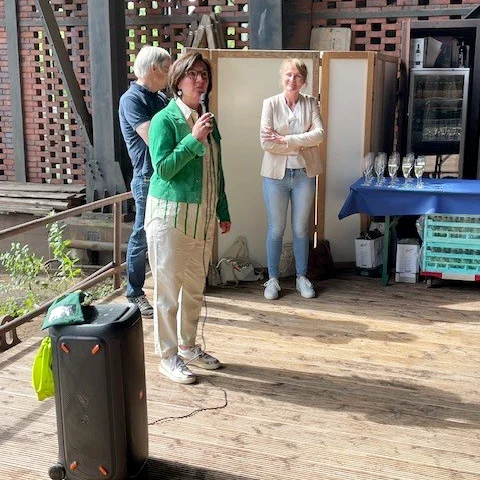 Das Foto zeigt drei Personen, die auf einer Holzterrasse stehen, mit einer Ziegelwand im Hintergrund. Die Person links trägt eine grüne Jacke und Jeans und hält ein Mikrofon in der Nähe ihres Mundes, um zu sprechen. Die Person rechts trägt ein weißes Oberteil und helle Hosen und hat die Hände vor sich verschränkt. Neben der Person links steht ein schwarzer Lautsprecher.Hinter ihnen befinden sich Regale mit verschiedenen Gegenständen und was wie Glaswaren aussieht.