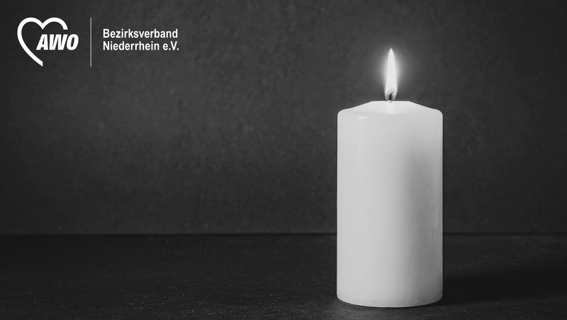 Auf dem Bild ist eine brennende, weiße Kerze auf dunklem Hintergrund zu sehen, insgesamt in Schwarz-Weiß gehalten. Links oben steht das AWO-Logo mit dem Text „Bezirksverband Niederrhein e.V.“, sodass das Motiv wie eine schlichte Trauer- oder Gedenkgrafik wirkt.