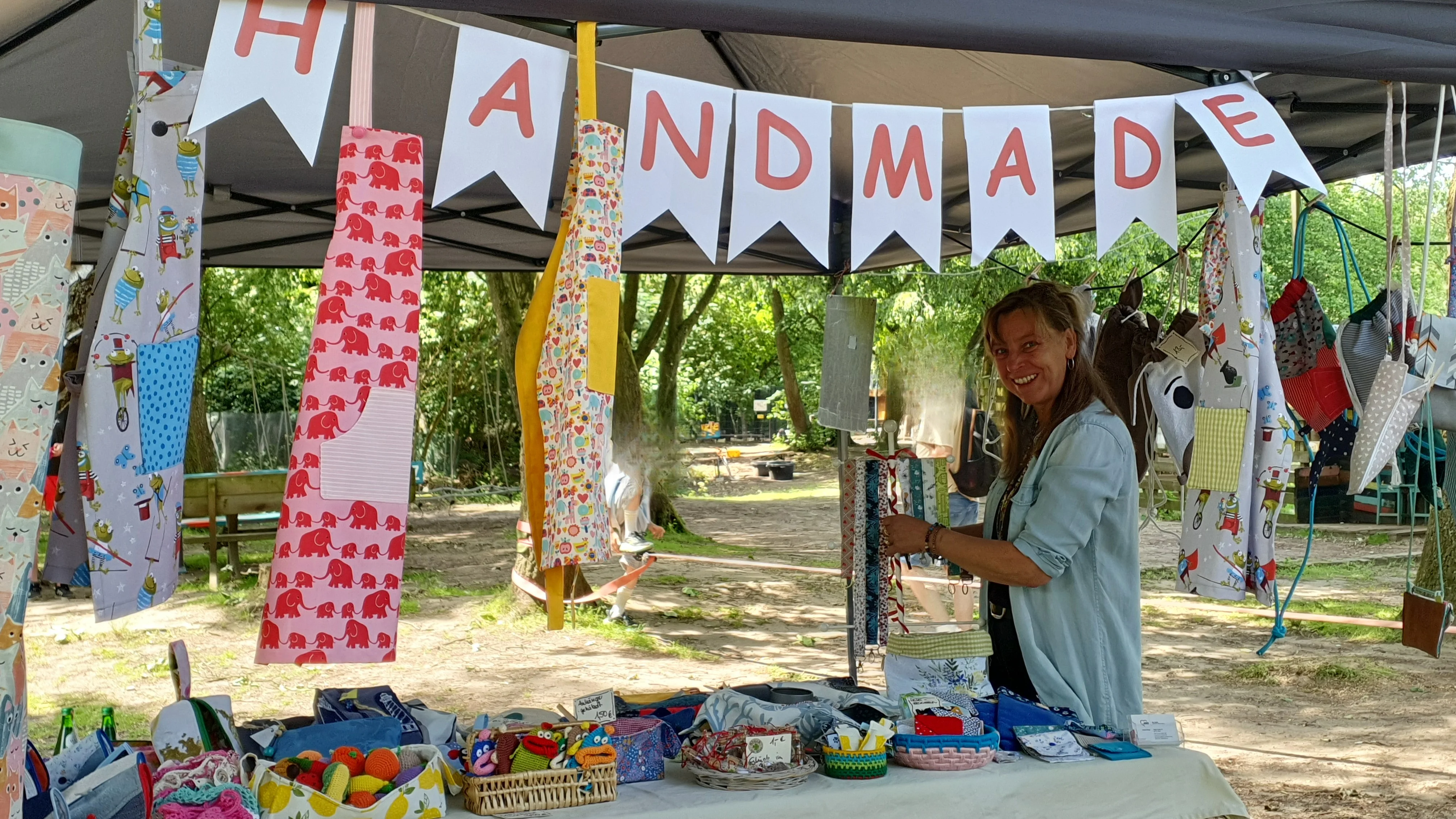 Auf dem Bild ist ein Markt- bzw. Infostand im Freien zu sehen, der unter einem Pavillon aufgebaut ist. Vorne steht ein Tisch mit vielen selbstgemachten Produkten, zum Beispiel genähten oder gehäkelten Artikeln, Täschchen und kleinen bunten Accessoires. Über dem Stand hängt eine Wimpelkette mit der Aufschrift „HANDMADE“. Rechts im Bild steht eine Person hinter dem Stand und ordnet oder präsentiert die Ware; im Hintergrund erkennt man Bäume, Spiel- oder Sitzbereich.