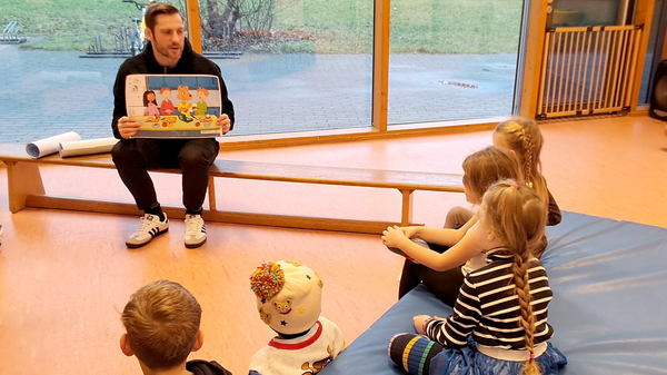 Auf dem Bild ist eine pädagogische Aktivität in einer Einrichtung zu sehen. Konkret erkennt man: Einen erwachsenen Mann in schwarzer Kleidung und Adidas-Schuhen, der auf einer Bank vor großen Fenstern sitzt und ein Bilderbuch mit bunten Cartoon-Figuren hochhält Mehrere Kinder (etwa 4-5), die auf einer blauen Turnmatte auf dem Boden sitzen und dem Vorleser zuhören Die Szene spielt in einem Raum mit hellem Holzinterieur, orangen/peachfarbenen Wänden und großen Fenstern mit Blick nach draußen Im Hintergrund sind eine Wiese und ein Gehweg zu sehen Die Atmosphäre wirkt entspannt und typisch für eine Vorlesestunde in einer pädagogischen Einrichtung