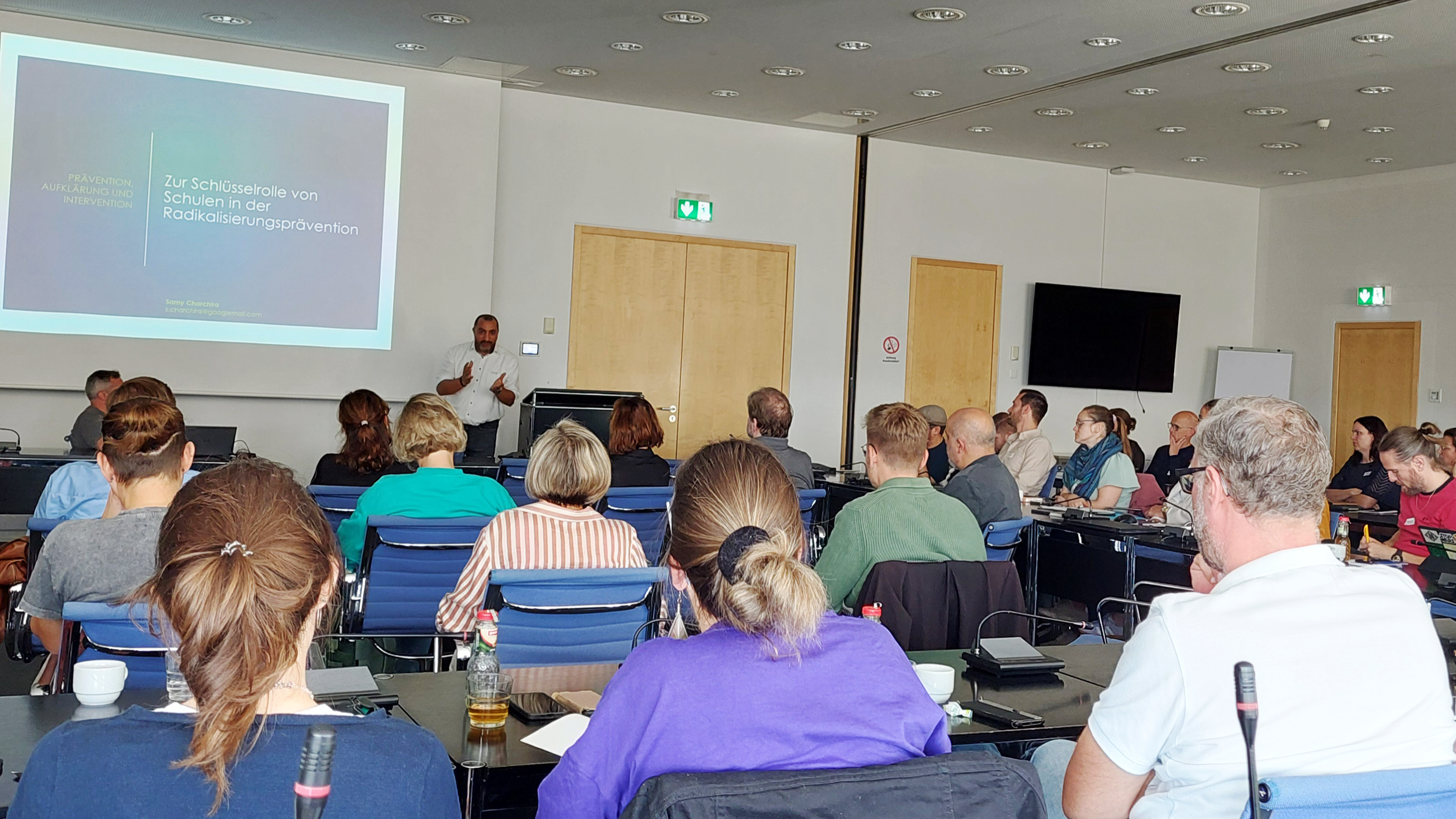 Auf dem Foto ist eine Gruppe von Personen in einem Konferenzraum zu sehen. Sie sitzen und schauen auf einen Präsentator, der vorne neben einer Projektionsleinwand steht. Die Leinwand zeigt den Text "Zur Schlüsselrolle von Schulen in der Radikalisierungsprävention". 
