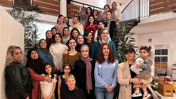 Auf dem Foto ist eine große Gruppe von Frauen und einigen jungen Mädchen zu sehen. Sie stehen und sitzen auf einer Treppe und im Erdgeschoss eines Innenraumes. Der Raum wirkt wie ein Gemeinschaftsraum oder ein Flur mit einer ansprechenden Treppe. Die Atmosphäre scheint freundlich und gemeinschaftlich zu sein. Einige Details, die man erkennen kann: Personen: Es sind etwa 25-30 Frauen und einige Mädchen unterschiedlichen Alters abgebildet. Kleidung: Die Kleidung ist vielfältig; einige Frauen tragen Kopftücher oder Hijabs, andere nicht. Der Stil reicht von leger bis etwas formeller. Hintergrund: Man sieht eine Treppe mit einem Metallgeländer, Pflanzen, eine helle Wand und eine Decke mit einer großen, runden Beleuchtung. Rechts im Bild ist ein Tisch mit einigen Kisten und Dekorationen zu erkennen, was auf eine Veranstaltung oder ein Treffen hindeutet.