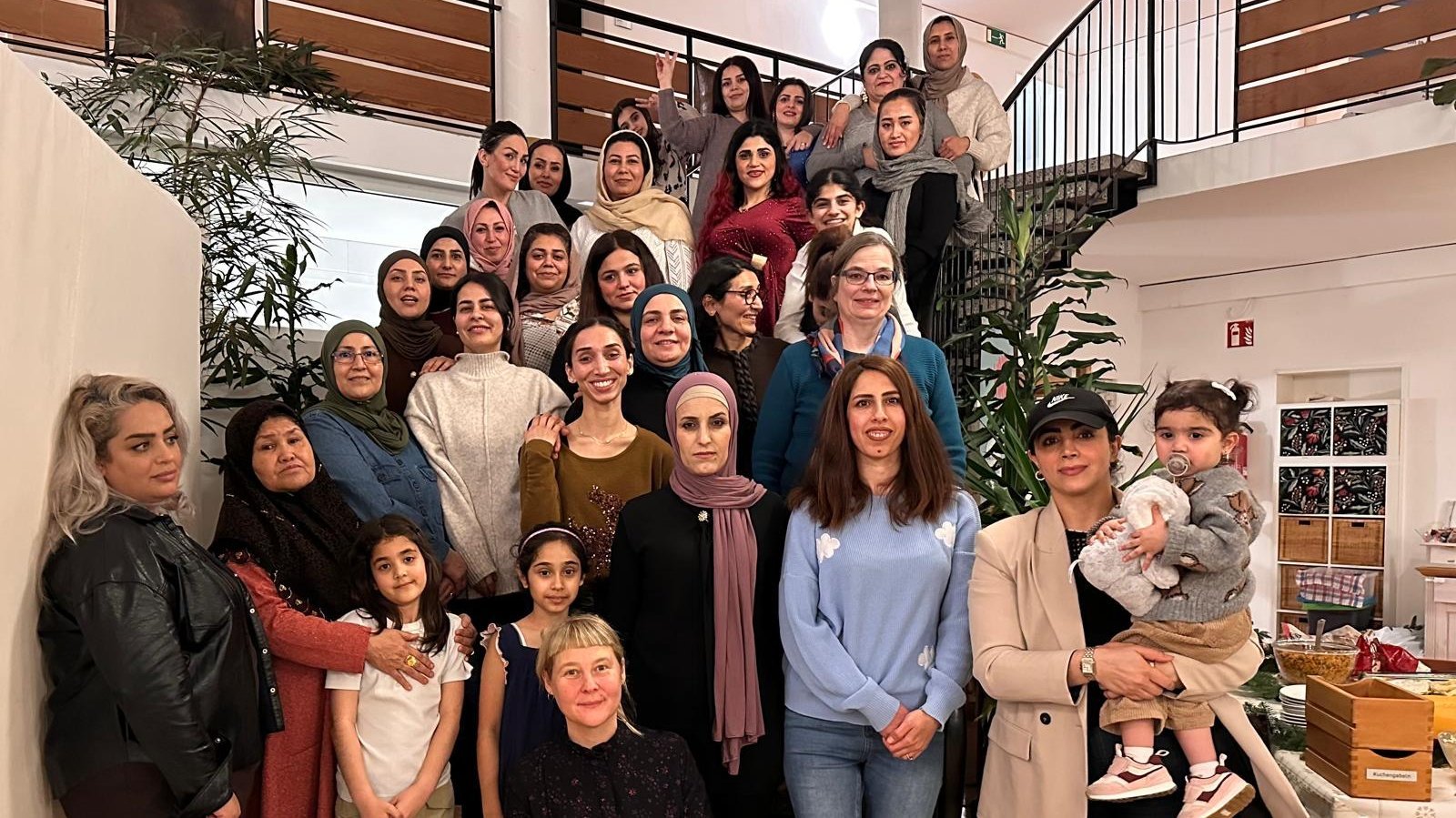 Auf dem Foto ist eine große Gruppe von Frauen und einigen jungen Mädchen zu sehen. Sie stehen und sitzen auf einer Treppe und im Erdgeschoss eines Innenraumes. Der Raum wirkt wie ein Gemeinschaftsraum oder ein Flur mit einer ansprechenden Treppe. Die Atmosphäre scheint freundlich und gemeinschaftlich zu sein. Einige Details, die man erkennen kann: Personen: Es sind etwa 25-30 Frauen und einige Mädchen unterschiedlichen Alters abgebildet. Kleidung: Die Kleidung ist vielfältig; einige Frauen tragen Kopftücher oder Hijabs, andere nicht. Der Stil reicht von leger bis etwas formeller. Hintergrund: Man sieht eine Treppe mit einem Metallgeländer, Pflanzen, eine helle Wand und eine Decke mit einer großen, runden Beleuchtung. Rechts im Bild ist ein Tisch mit einigen Kisten und Dekorationen zu erkennen, was auf eine Veranstaltung oder ein Treffen hindeutet.