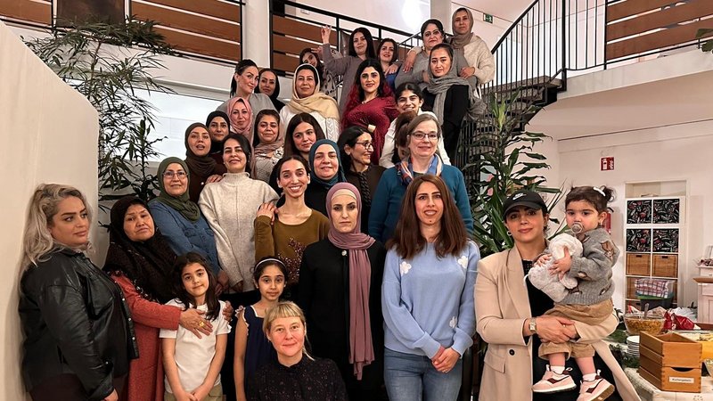 Auf dem Foto ist eine große Gruppe von Frauen und einigen jungen Mädchen zu sehen. Sie stehen und sitzen auf einer Treppe und im Erdgeschoss eines Innenraumes. Der Raum wirkt wie ein Gemeinschaftsraum oder ein Flur mit einer ansprechenden Treppe. Die Atmosphäre scheint freundlich und gemeinschaftlich zu sein. Einige Details, die man erkennen kann: Personen: Es sind etwa 25-30 Frauen und einige Mädchen unterschiedlichen Alters abgebildet. Kleidung: Die Kleidung ist vielfältig; einige Frauen tragen Kopftücher oder Hijabs, andere nicht. Der Stil reicht von leger bis etwas formeller. Hintergrund: Man sieht eine Treppe mit einem Metallgeländer, Pflanzen, eine helle Wand und eine Decke mit einer großen, runden Beleuchtung. Rechts im Bild ist ein Tisch mit einigen Kisten und Dekorationen zu erkennen, was auf eine Veranstaltung oder ein Treffen hindeutet.