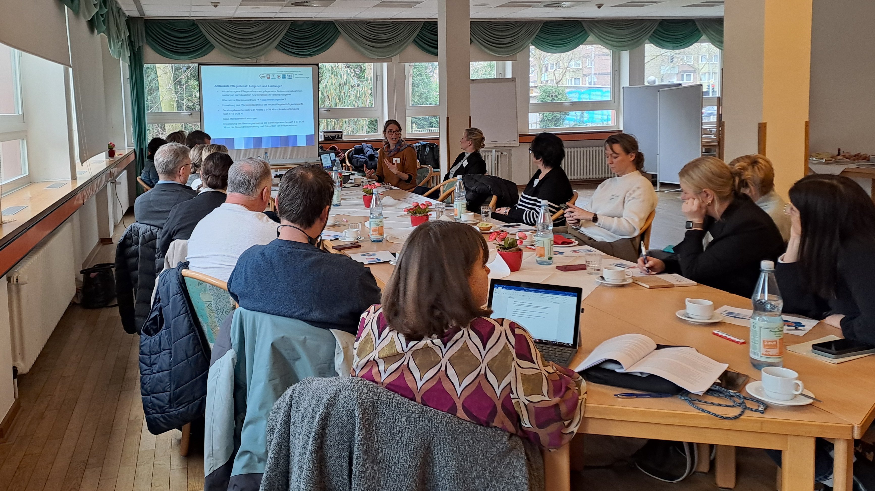 Auf dem Foto ist eine größere Besprechung oder Tagung in einem Saal zu sehen. Die Personen sitzen an einem U‑förmig gestellten Tisch, haben Laptops, Unterlagen, Getränke und Kaffeetassen vor sich und schauen überwiegend nach vorne auf eine Leinwand, auf der eine Präsentation projiziert wird.