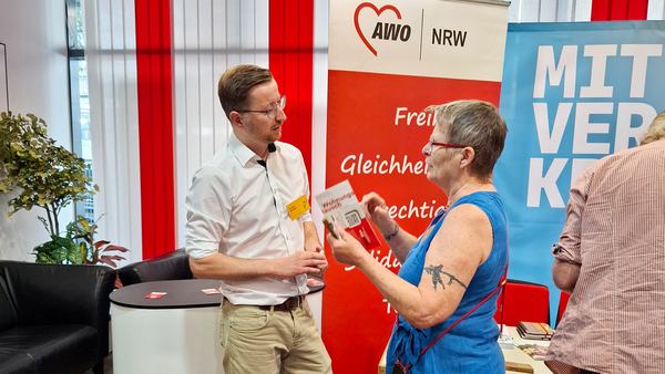 Auf dem Foto ist eine Szene von einer Veranstaltung mit der AWO NRW zu sehen: • Im Vordergrund unterhalten sich ein Mann und eine Frau angeregt miteinander. o Der Mann trägt ein weißes Hemd und ein Namensschild. o Die Frau trägt ein ärmelloses blaues Kleid, rote Schuhe und hält einen AWO-Flyer in der Hand. Sie hat eine auffällige Tätowierung am Oberarm. • Im Hintergrund sind Roll-Ups der AWO NRW zu sehen mit den Worten „Freiheit, Gleichheit, Gerechtigkeit, Solidarität“ und ein weiteres mit dem Schriftzug „MIT VER...“ (vermutlich „MITVERANTWORTUNG“ oder „MITVERÄNDERUNG“). • Es handelt sich offenbar um einen Informations- oder Austauschstand der AWO NRW in einem Innenraum mit moderner Büroeinrichtung (Sitzecke, Schreibtisch, rote Vorhänge). Die Szene vermittelt den Eindruck eines offenen Dialogs zwischen AWO-Mitarbeitenden und Interessierten im Rahmen einer Infoveranstaltung.