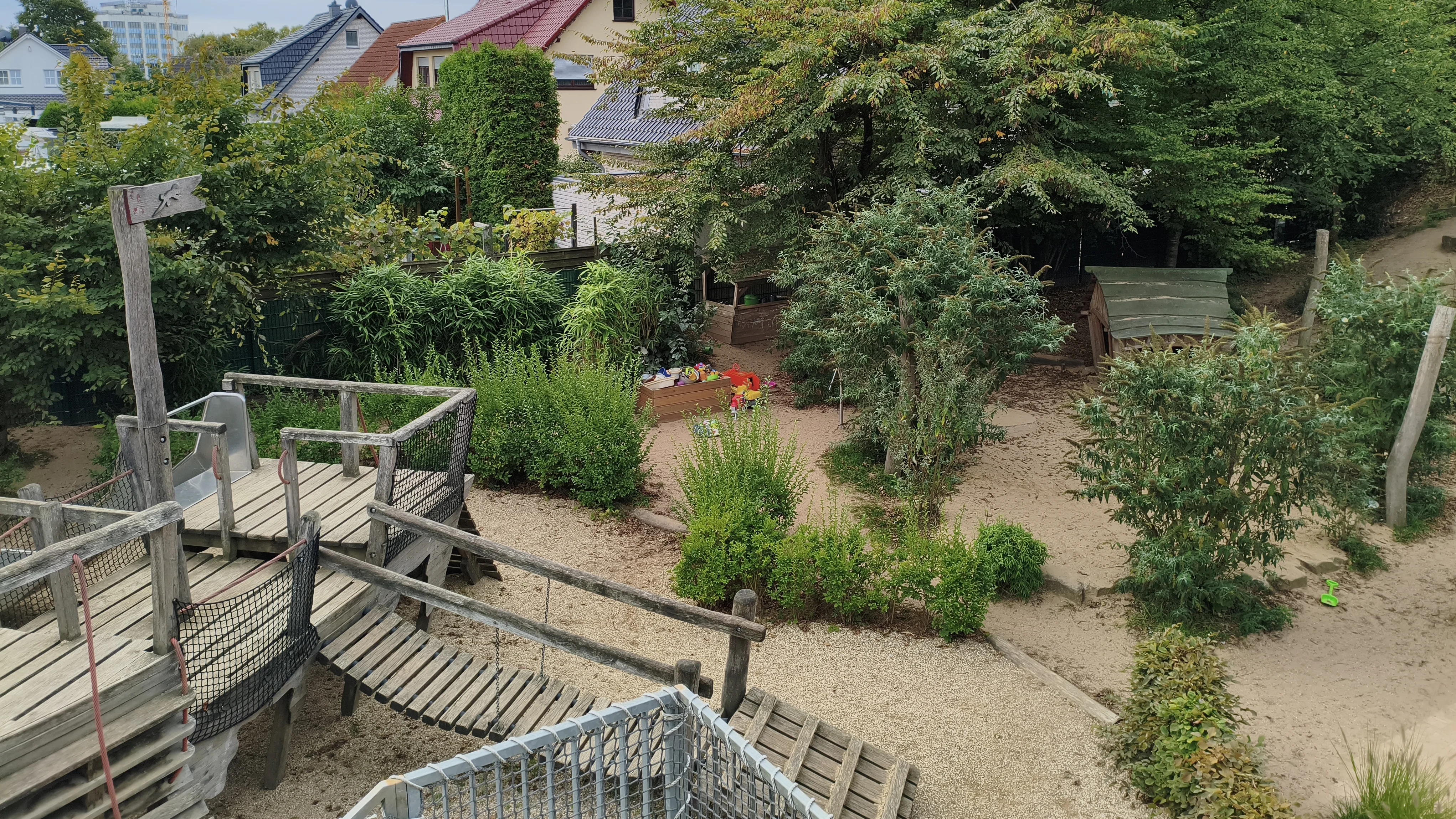 Auf dem Foto ist ein Spielplatz zu sehen, der in einer Wohngegend liegt und von viel Grün umgeben ist. Hier sind die Details, die man erkennen kann: Vordergrund (links unten): Ein hölzernes Spielgerät mit einer Rutsche, einer Hängebrücke und Treppen oder Aufgängen. Spielbereich: Der Boden des Spielplatzes besteht hauptsächlich aus Sand oder feinem Kies. Mitte: Es gibt Büsche und kleinere Bäume, die den Bereich natürlich gliedern. In der Mitte, etwas zurückgesetzt, ist ein kleiner Sandkasten mit einigen bunten Spielsachen (Schaufeln, Eimer) zu erkennen. Hintergrund: Man sieht die Dächer von Wohnhäusern (mit roten Ziegeln), was darauf hindeutet, dass der Spielplatz in einer Siedlung oder einem Wohngebiet liegt. Umgebung: Der Spielplatz ist von vielen Bäumen und Sträuchern umgeben, die ihm eine natürliche, fast waldartige Atmosphäre verleihen.