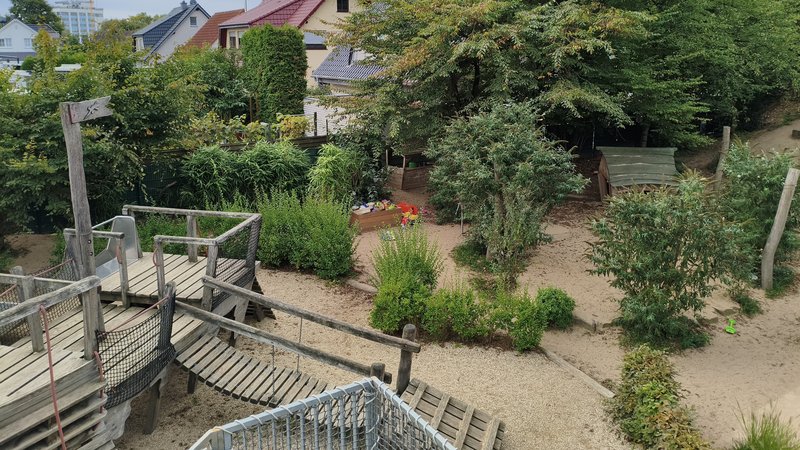 Auf dem Foto ist ein Spielplatz zu sehen, der in einer Wohngegend liegt und von viel Grün umgeben ist. Hier sind die Details, die man erkennen kann: Vordergrund (links unten): Ein hölzernes Spielgerät mit einer Rutsche, einer Hängebrücke und Treppen oder Aufgängen. Spielbereich: Der Boden des Spielplatzes besteht hauptsächlich aus Sand oder feinem Kies. Mitte: Es gibt Büsche und kleinere Bäume, die den Bereich natürlich gliedern. In der Mitte, etwas zurückgesetzt, ist ein kleiner Sandkasten mit einigen bunten Spielsachen (Schaufeln, Eimer) zu erkennen. Hintergrund: Man sieht die Dächer von Wohnhäusern (mit roten Ziegeln), was darauf hindeutet, dass der Spielplatz in einer Siedlung oder einem Wohngebiet liegt. Umgebung: Der Spielplatz ist von vielen Bäumen und Sträuchern umgeben, die ihm eine natürliche, fast waldartige Atmosphäre verleihen.