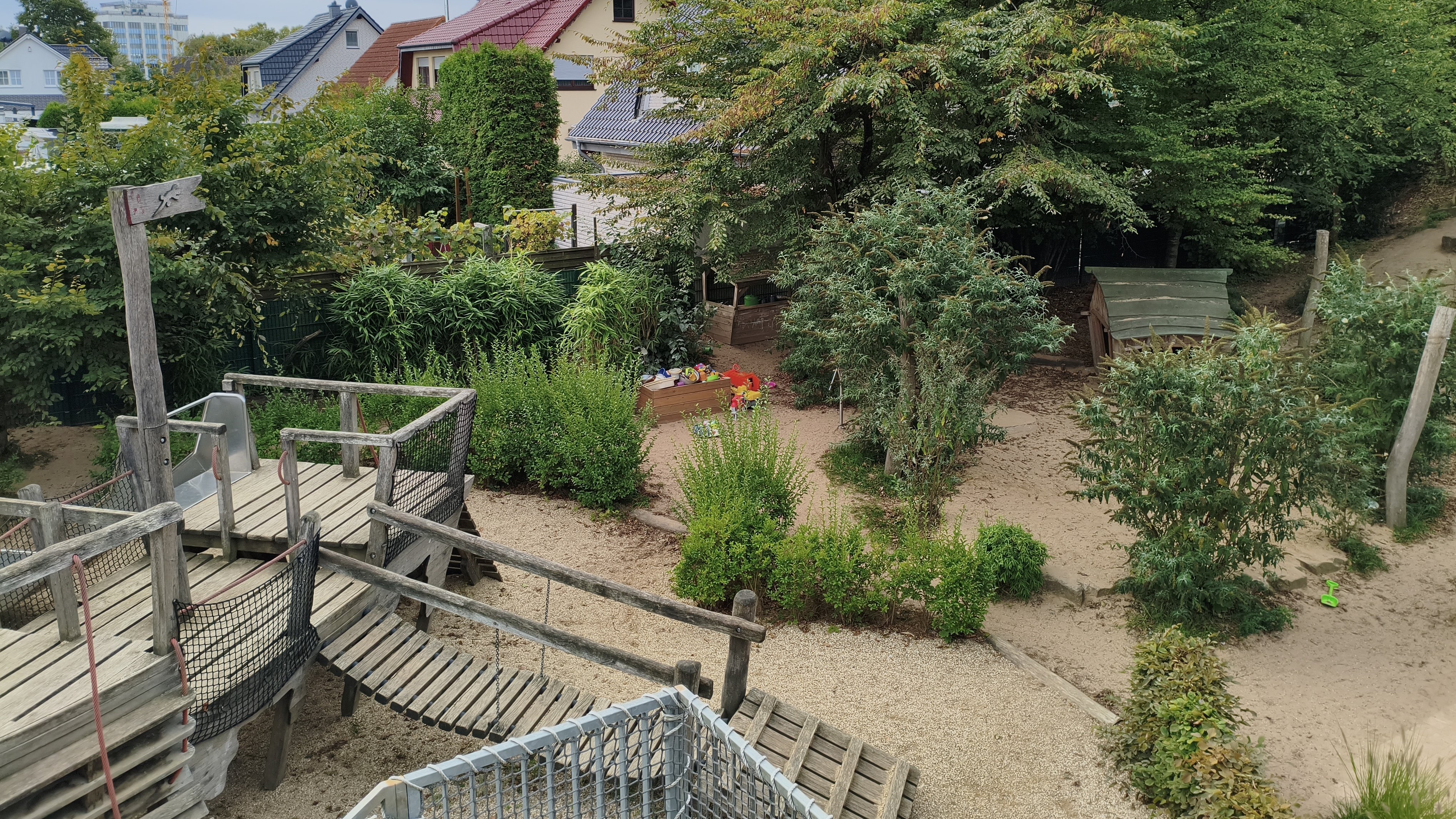 Auf dem Foto ist ein Spielplatz zu sehen, der in einer Wohngegend liegt und von viel Grün umgeben ist. Hier sind die Details, die man erkennen kann: Vordergrund (links unten): Ein hölzernes Spielgerät mit einer Rutsche, einer Hängebrücke und Treppen oder Aufgängen. Spielbereich: Der Boden des Spielplatzes besteht hauptsächlich aus Sand oder feinem Kies. Mitte: Es gibt Büsche und kleinere Bäume, die den Bereich natürlich gliedern. In der Mitte, etwas zurückgesetzt, ist ein kleiner Sandkasten mit einigen bunten Spielsachen (Schaufeln, Eimer) zu erkennen. Hintergrund: Man sieht die Dächer von Wohnhäusern (mit roten Ziegeln), was darauf hindeutet, dass der Spielplatz in einer Siedlung oder einem Wohngebiet liegt. Umgebung: Der Spielplatz ist von vielen Bäumen und Sträuchern umgeben, die ihm eine natürliche, fast waldartige Atmosphäre verleihen.