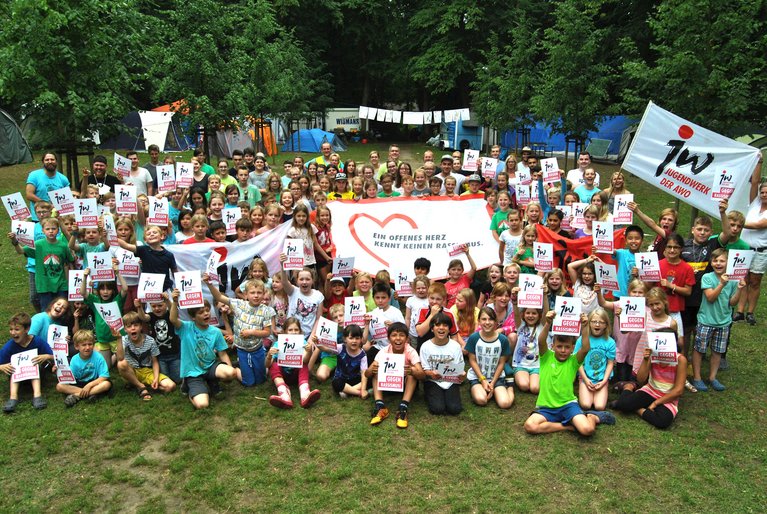 Foto Zeltlager Elten: Zu sehen sind etwa 100 Kinder, die auf einer Wiese stehen. Viele halten Plakate mit der Aufschrift "Jugendwerk gegen Rassismus" in die Höhe. In der Mitte der Gruppe eine große Fahne mit dem Aufgruck "Ein offenes Herz kennen keinen Rassismus". Im Hintergrund sind viele Zelte zu sehen.