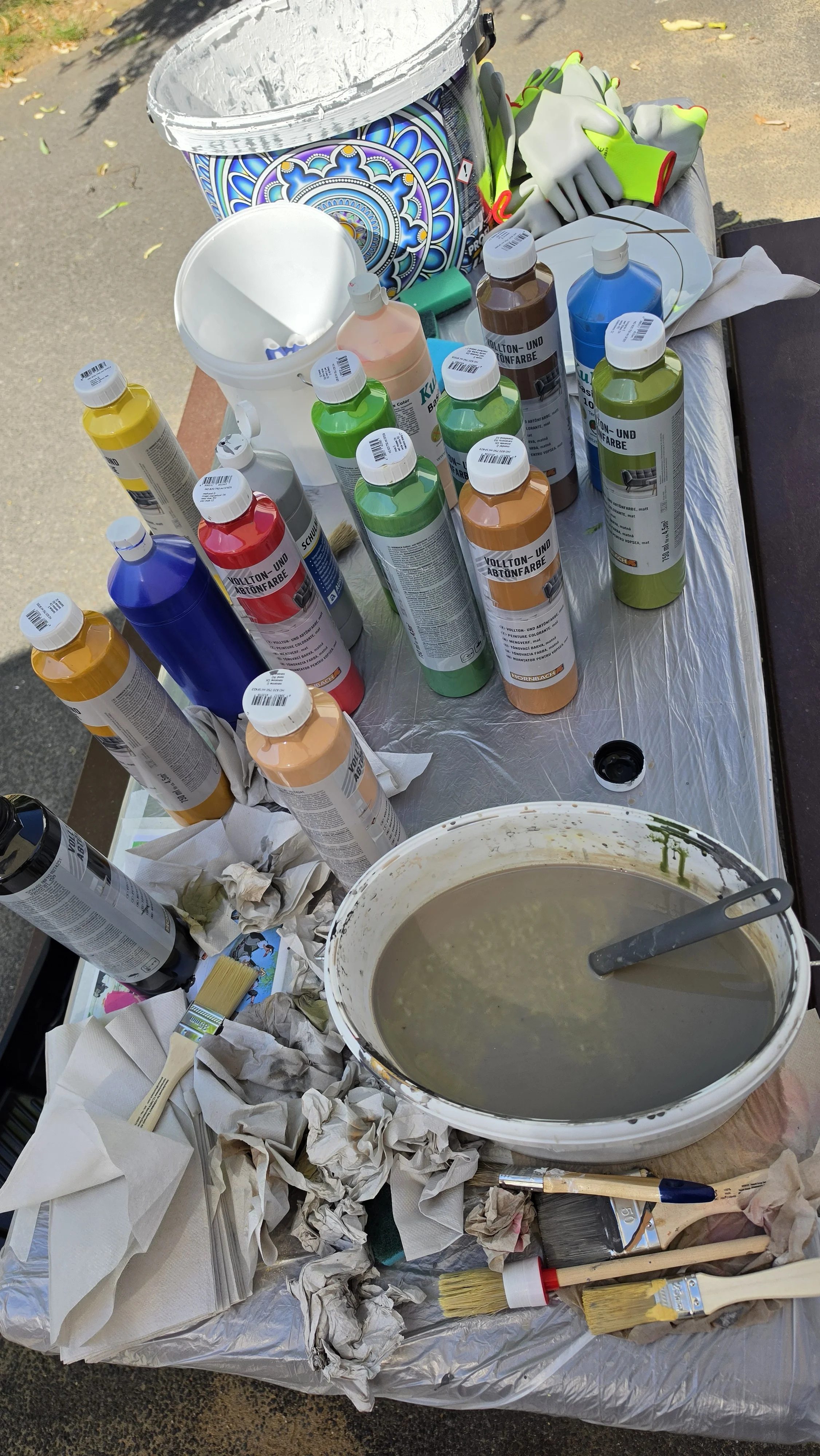 Auf dem Bild ist ein Arbeitstisch im Freien zu sehen, der für Mal‑ und Streicharbeiten vorbereitet ist. Auf dem Tisch stehen viele Flaschen mit bunten Acryl‑ oder Abtönfarben in verschiedenen Farben (Gelb, Blau, Rot, Grün, Braun usw.), daneben Eimer und Schalen mit Wasser bzw. verdünnter Farbe. Vorne liegen Pinsel und zusammengeknüllte Papiertücher, im Hintergrund erkennt man einen großen Farbkübel mit dekorativem Muster sowie Arbeitshandschuhe – insgesamt also ein kompletter Maler‑/Graffiti‑Arbeitsplatz für ein kreatives Projekt.