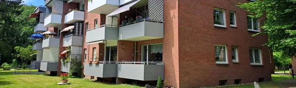 Auf dem Foto ist ein mehrstöckiges Wohnhaus zu sehen, vermutlich ein Mehrfamilienhaus. Es besteht aus rotem Backstein mit modernen, grau-weißen Balkonen, die bei vielen Wohnungen vorhanden sind. Einige Balkone sind mit Pflanzen, Möbeln oder Sonnenschirmen geschmückt. Das Gebäude hat drei Etagen, und vor dem Haus befindet sich eine gepflegte Rasenfläche mit einem einzelnen Blumenkübel sowie einer Sitzbank. Im Hintergrund ist viel Grün zu sehen, was auf eine ruhige und naturnahe Umgebung hindeutet. Das Wetter ist sonnig mit blauem Himmel und ein paar Wolken.