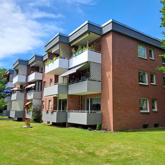 Auf dem Foto ist ein mehrstöckiges Wohnhaus zu sehen, vermutlich ein Mehrfamilienhaus. Es besteht aus rotem Backstein mit modernen, grau-weißen Balkonen, die bei vielen Wohnungen vorhanden sind. Einige Balkone sind mit Pflanzen, Möbeln oder Sonnenschirmen geschmückt. Das Gebäude hat drei Etagen, und vor dem Haus befindet sich eine gepflegte Rasenfläche mit einem einzelnen Blumenkübel sowie einer Sitzbank. Im Hintergrund ist viel Grün zu sehen, was auf eine ruhige und naturnahe Umgebung hindeutet. Das Wetter ist sonnig mit blauem Himmel und ein paar Wolken.