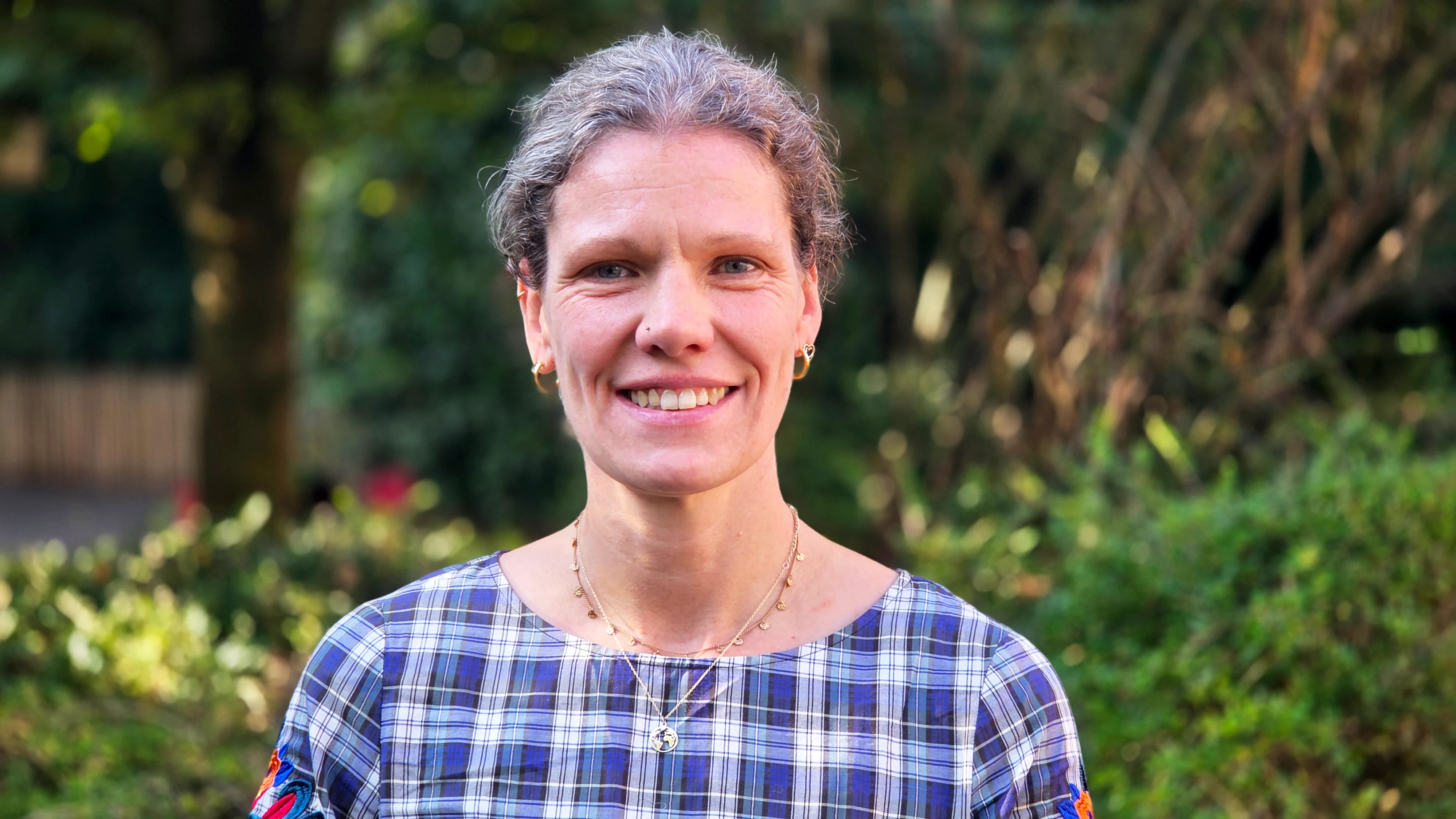 Auf dem Bild ist eine lächelnde Frau als Porträt zu sehen, vor einem unscharfen grünen Hintergrund im Freien. Sie trägt ein blau-weiß kariertes Oberteil und Schmuck an den Ohren und am Hals.