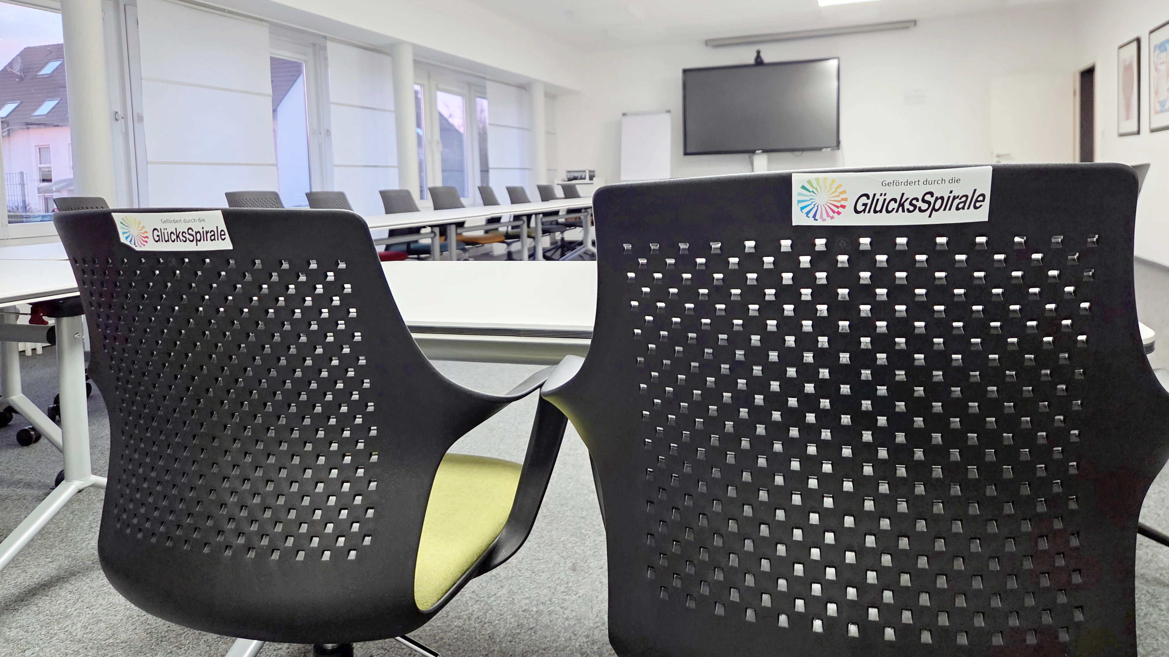 Auf dem Bild ist ein moderner Konferenz- oder Schulungsraum zu sehen, der augenscheinlich neu ausgestattet wurde. Hier sind die Details: Vordergrund: Im Vordergrund sind zwei ergonomisch gestaltete Stühle mit dunklen, perforierten Rückenlehnen zu sehen. Auf beiden Stühlen ist ein Aufkleber mit dem Logo und dem Text "Gefördert durch die GlücksSpirale" angebracht, was auf die Finanzierung der neuen Ausstattung hinweist. Möbel: Im Hintergrund sind weitere Tische und Stühle erkennbar, die in einer U- oder Blockform angeordnet sind. Die Tische scheinen modular und flexibel zu sein, was zu der Beschreibung einer modernen Konferenzausstattung passt. Technik: An der Stirnseite des Raumes ist ein großer Flachbildschirm (vermutlich für Präsentationen oder Videokonferenzen) an der Wand montiert. Raumgestaltung: Der Raum wirkt hell und aufgeräumt mit einer abgehängten Decke, in die quadratische LED-Leuchten eingelassen sind. Die Fensterfront ist mit hellen Rollos oder Jalousien versehen. Gesamteindruck: Das Bild vermittelt den Eindruck eines professionellen, zeitgemäßen Tagungsortes, der für Schulungen, Besprechungen oder Konferenzen eingerichtet wurde.