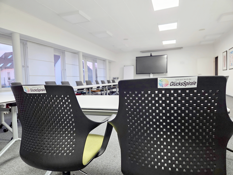 Auf dem Bild ist ein moderner Konferenz- oder Schulungsraum zu sehen, der augenscheinlich neu ausgestattet wurde. Hier sind die Details: Vordergrund: Im Vordergrund sind zwei ergonomisch gestaltete Stühle mit dunklen, perforierten Rückenlehnen zu sehen. Auf beiden Stühlen ist ein Aufkleber mit dem Logo und dem Text "Gefördert durch die GlücksSpirale" angebracht, was auf die Finanzierung der neuen Ausstattung hinweist. Möbel: Im Hintergrund sind weitere Tische und Stühle erkennbar, die in einer U- oder Blockform angeordnet sind. Die Tische scheinen modular und flexibel zu sein, was zu der Beschreibung einer modernen Konferenzausstattung passt. Technik: An der Stirnseite des Raumes ist ein großer Flachbildschirm (vermutlich für Präsentationen oder Videokonferenzen) an der Wand montiert. Raumgestaltung: Der Raum wirkt hell und aufgeräumt mit einer abgehängten Decke, in die quadratische LED-Leuchten eingelassen sind. Die Fensterfront ist mit hellen Rollos oder Jalousien versehen. Gesamteindruck: Das Bild vermittelt den Eindruck eines professionellen, zeitgemäßen Tagungsortes, der für Schulungen, Besprechungen oder Konferenzen eingerichtet wurde.
