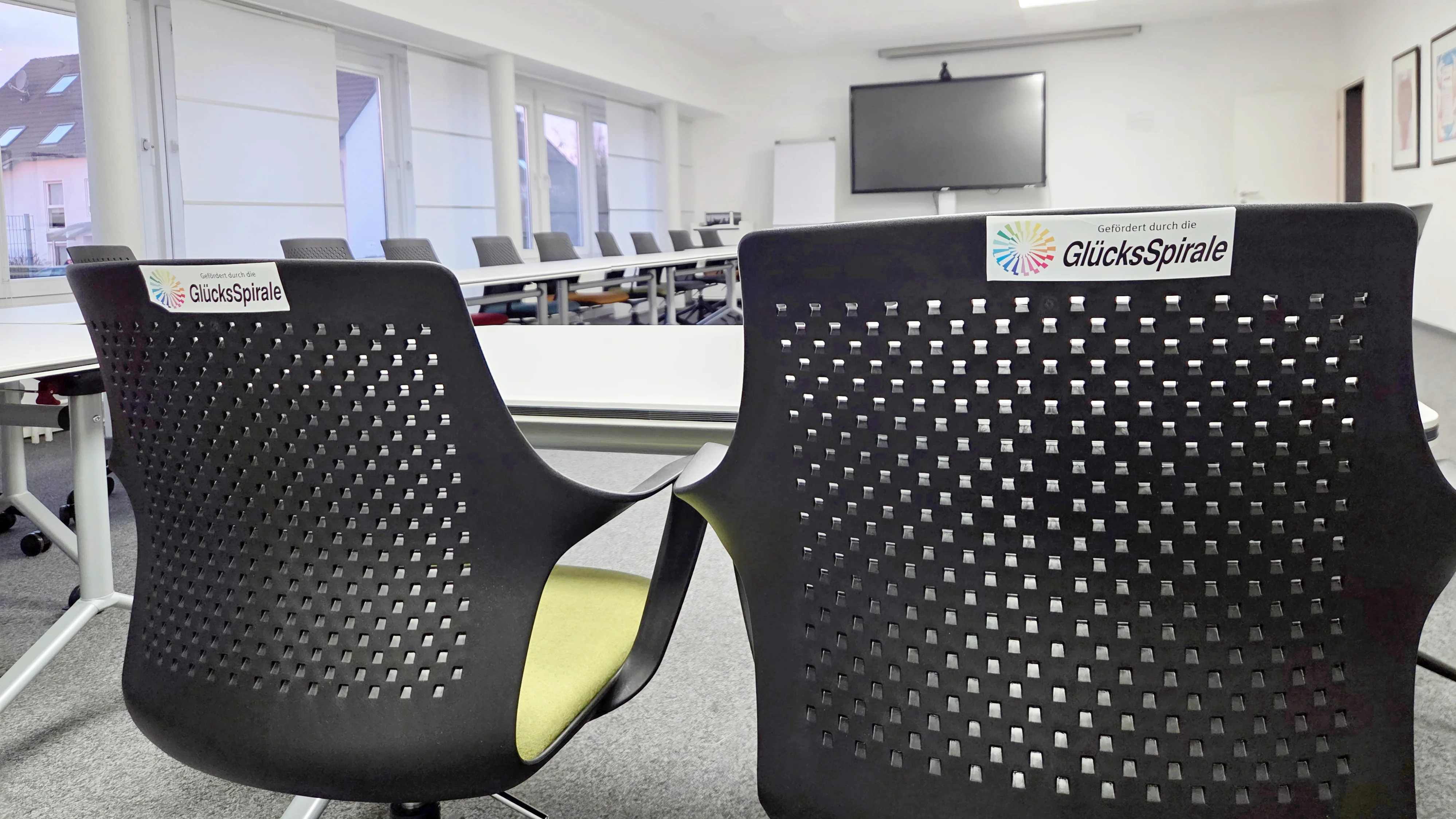 Auf dem Bild ist ein moderner Konferenz- oder Schulungsraum zu sehen, der augenscheinlich neu ausgestattet wurde. Hier sind die Details: Vordergrund: Im Vordergrund sind zwei ergonomisch gestaltete Stühle mit dunklen, perforierten Rückenlehnen zu sehen. Auf beiden Stühlen ist ein Aufkleber mit dem Logo und dem Text "Gefördert durch die GlücksSpirale" angebracht, was auf die Finanzierung der neuen Ausstattung hinweist. Möbel: Im Hintergrund sind weitere Tische und Stühle erkennbar, die in einer U- oder Blockform angeordnet sind. Die Tische scheinen modular und flexibel zu sein, was zu der Beschreibung einer modernen Konferenzausstattung passt. Technik: An der Stirnseite des Raumes ist ein großer Flachbildschirm (vermutlich für Präsentationen oder Videokonferenzen) an der Wand montiert. Raumgestaltung: Der Raum wirkt hell und aufgeräumt mit einer abgehängten Decke, in die quadratische LED-Leuchten eingelassen sind. Die Fensterfront ist mit hellen Rollos oder Jalousien versehen. Gesamteindruck: Das Bild vermittelt den Eindruck eines professionellen, zeitgemäßen Tagungsortes, der für Schulungen, Besprechungen oder Konferenzen eingerichtet wurde.