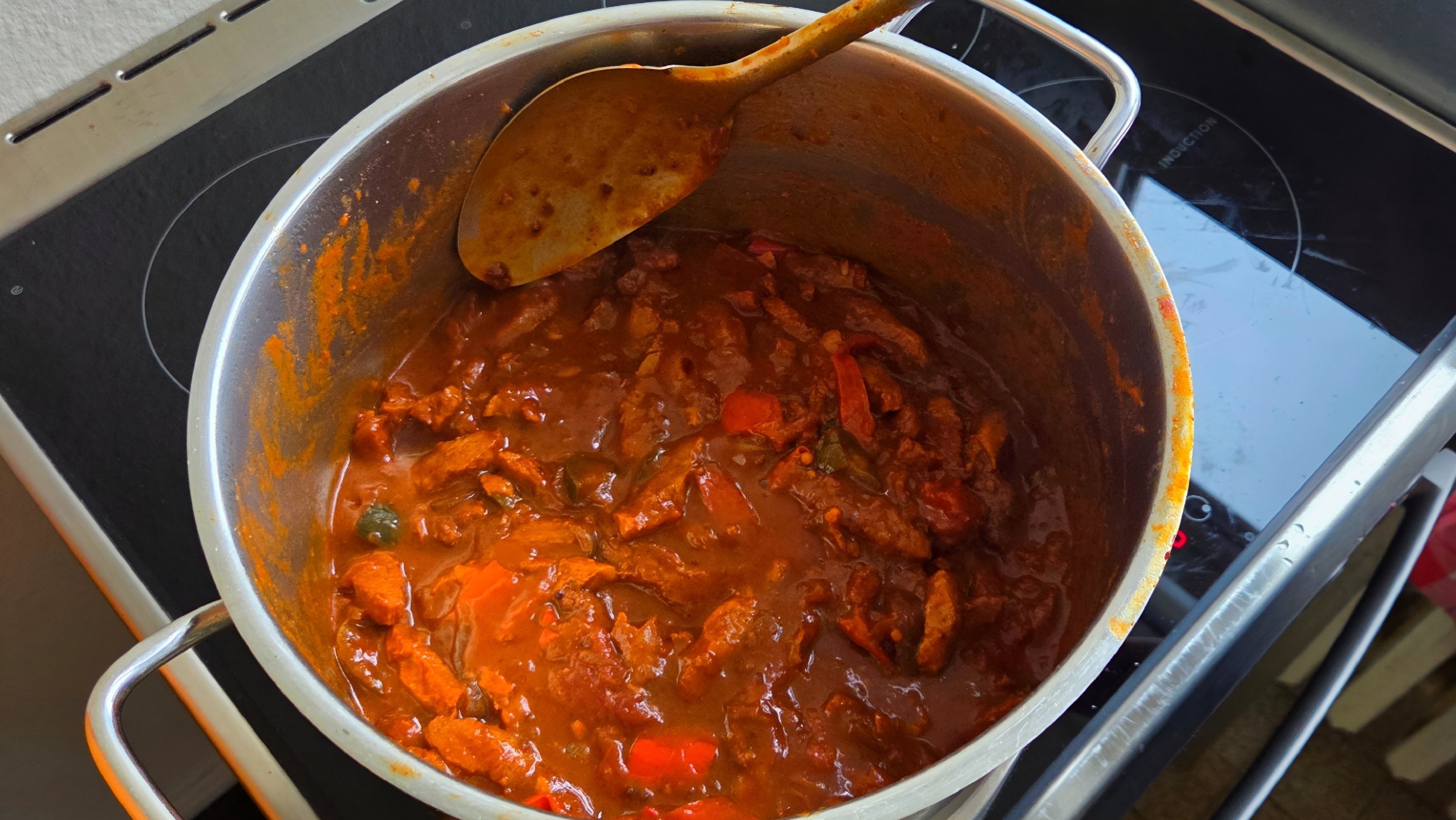 Auf dem Bild sieht man einen großen Kochtopf mit einer dickflüssigen, tomatigen Gulaschsoße mit Gemüsestücken, der auf einem Herd steht. Eine Hand hält einen Kochlöffel über dem Topf, daneben sind Teile der Küche wie Herdplatte, Bodenfliesen und ein Fensterrahmen zu erkennen.