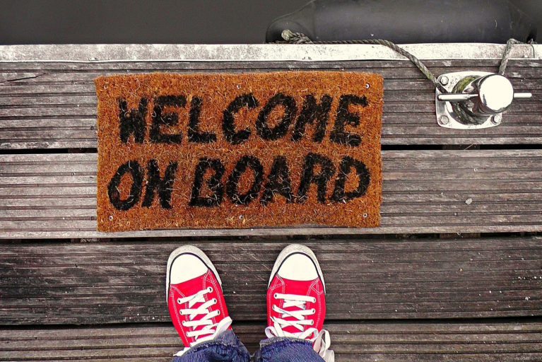 Das Foto zeigt Füße vor einer Fußmatte mit der Aufschrift "Welcome on board" (Bild von Mabel Amber, who will one day auf Pixabay).