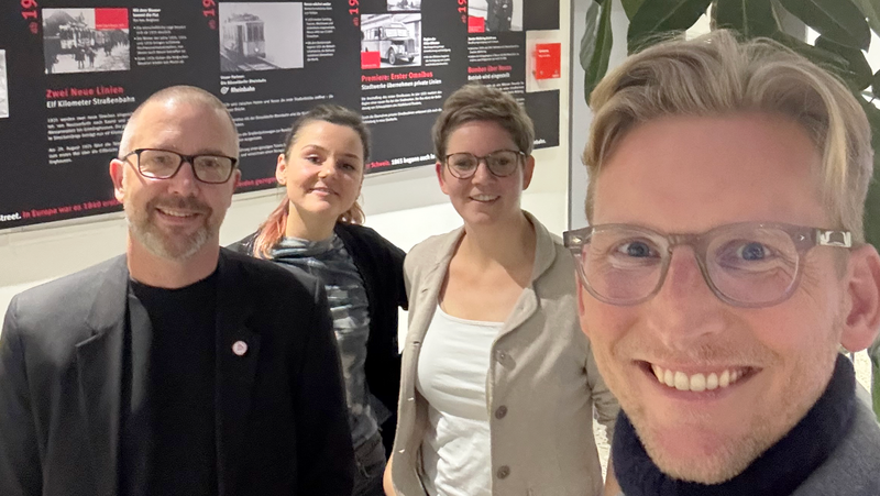 Auf dem Bild sind vier Personen zu sehen, die in einem Innenraum stehen und lächeln. Es sieht aus wie ein Gruppen-Selfie. Im Hintergrund befindet sich eine schwarz-rote Zeitleiste oder Ausstellungstafel mit Text und Bildern. Rechts im Bild steht außerdem eine große Grünpflanze.