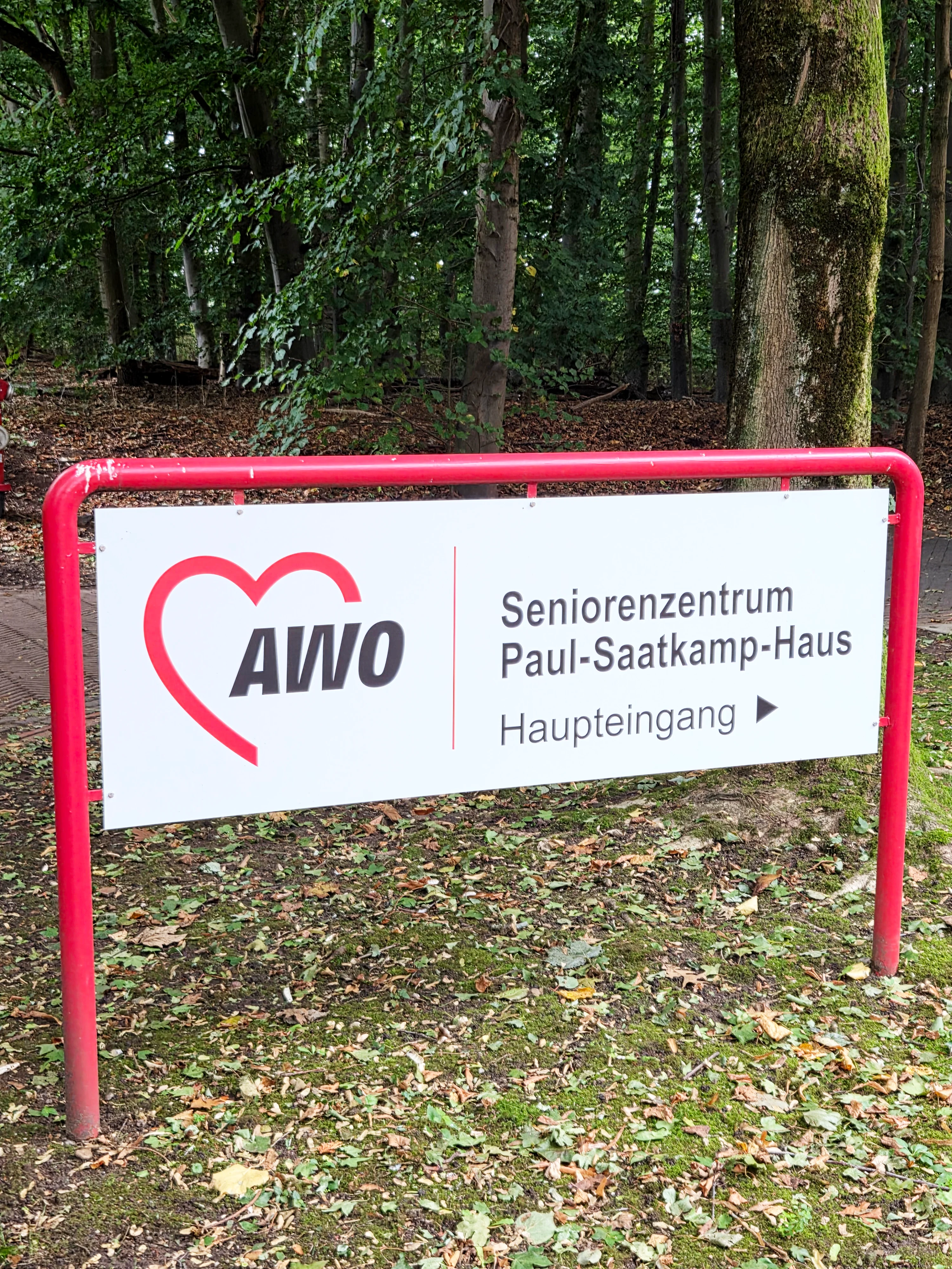 Auf dem Foto ist ein Schild zu sehen, das auf das "Seniorenzentrum Paul-Saatkamp-Haus" hinweist. Es hat einen weißen Hintergrund mit rotem Rahmen und roten Buchstaben. Auf der linken Seite des Schildes befindet sich das Logo der AWO (Arbeiterwohlfahrt), das aus einem stilisierten roten Herzen mit den Buchstaben "AWO" besteht. Auf der rechten Seite stehen die Worte "Seniorenzentrum Paul-Saatkamp-Haus" und darunter "Haupteingang" mit einem Pfeil, der nach rechts zeigt. Das Schild steht auf einem mit Blättern bedeckten Boden, und im Hintergrund sind Bäume zu erkennen, was darauf hindeutet, dass sich das Zentrum in einer grünen, waldähnlichen Umgebung befindet.