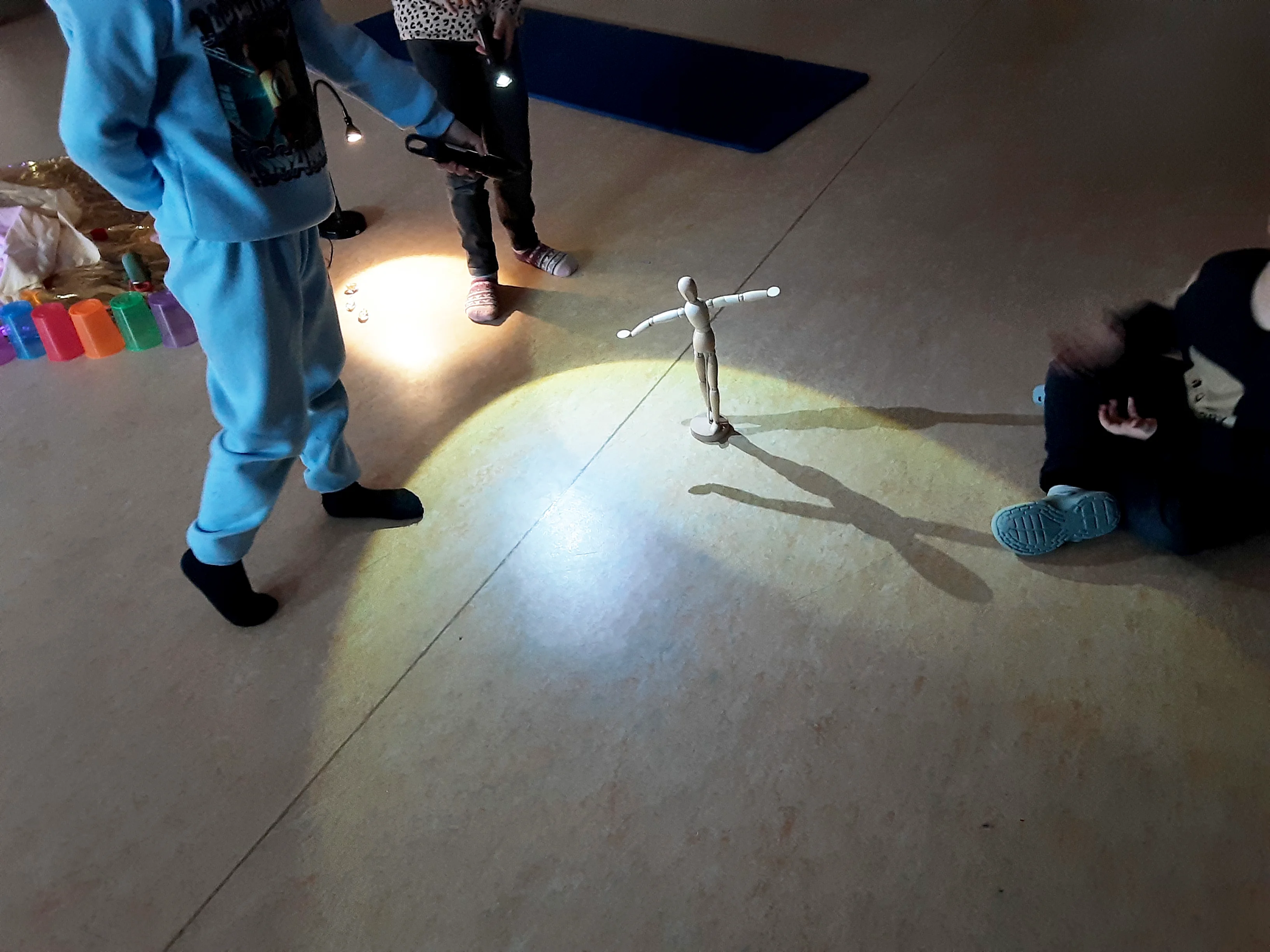 Auf dem Bild sind Kinder in einem Raum zu sehen, die mit Taschenlampen eine Holz‑Gliederpuppe anstrahlen. Die Puppe steht in der Mitte mit ausgebreiteten Armen und wirft durch das Licht deutlich sichtbare Schatten auf den Boden.