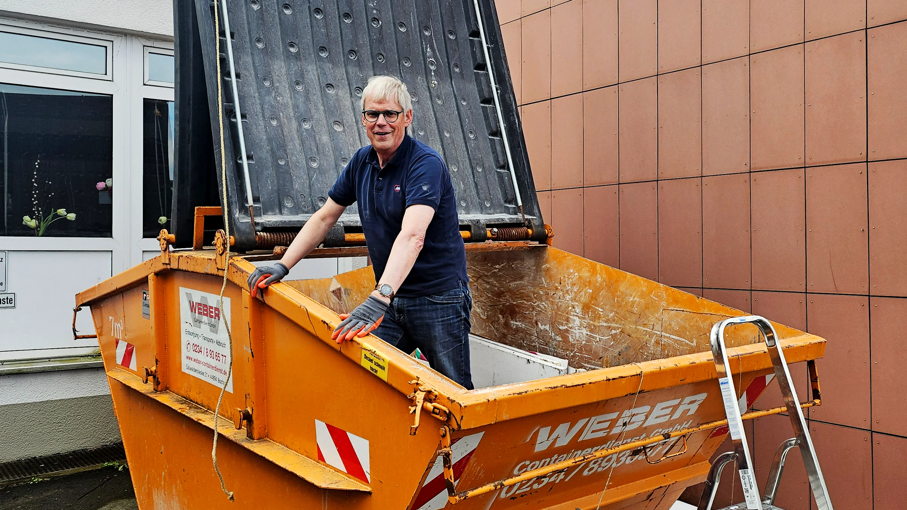 Das Foto zeigt eine Person, die eine dunkle Oberbekleidung und helle Hosen trägt. Sie steht in einem orangefarbenen Baumischcontainer. Die Person hält sich an den Geländern des Containers fest. Im Hintergrund ist ein Gebäude mit großen Fenstern zu sehen, die den Himmel und andere Gebäude reflektieren.