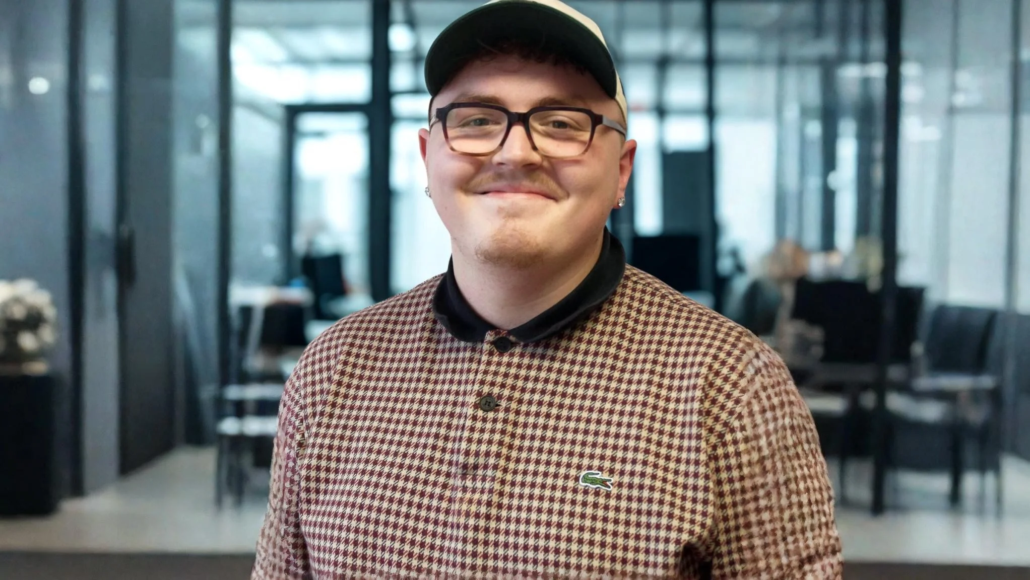 Auf dem Bild ist ein junger Mann zu sehen, der lächelnd in die Kamera schaut. Er trägt: eine schwarz-beige Snapback-Cap eine eckige, dunkle Brille ein kariertes Poloshirt/Pullover von Lacoste (bordeaux-beige Hahnentritt-Muster) mit schwarzem Kragen einen kleinen Ohrstecker Im Hintergrund ist ein modernes Büro- oder Empfangsgebäude mit Glaswänden zu erkennen, leicht unscharf (Bokeh-Effekt).