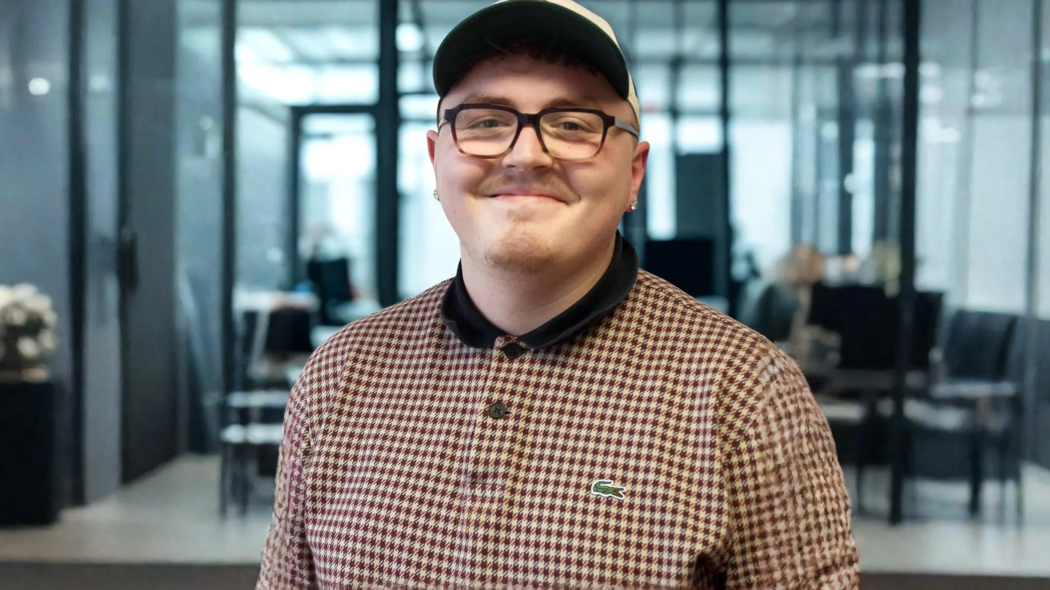 Auf dem Bild ist ein junger Mann zu sehen, der lächelnd in die Kamera schaut. Er trägt: eine schwarz-beige Snapback-Cap eine eckige, dunkle Brille ein kariertes Poloshirt/Pullover von Lacoste (bordeaux-beige Hahnentritt-Muster) mit schwarzem Kragen einen kleinen Ohrstecker Im Hintergrund ist ein modernes Büro- oder Empfangsgebäude mit Glaswänden zu erkennen, leicht unscharf (Bokeh-Effekt).