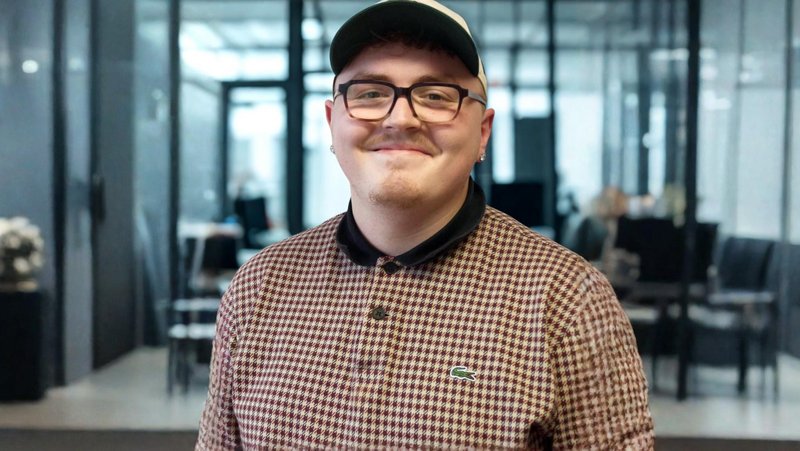 Auf dem Bild ist ein junger Mann zu sehen, der lächelnd in die Kamera schaut. Er trägt: ​ eine schwarz-beige Snapback-Cap eine eckige, dunkle Brille ein kariertes Poloshirt/Pullover von Lacoste (bordeaux-beige Hahnentritt-Muster) mit schwarzem Kragen einen kleinen Ohrstecker Im Hintergrund ist ein modernes Büro- oder Empfangsgebäude mit Glaswänden zu erkennen, leicht unscharf (Bokeh-Effekt).