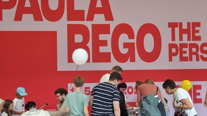 Auf dem Foto ist eine Gruppe von Leuten zu sehen, die in einer Kunstgalerie oder einem Museum zusammenkommen. Der Hintergrund zeigt eine große, rote Wand mit weißer Schrift, auf der „PAULA REGO“ und „THE PERSONAL“ zu lesen ist. Einige Leute sitzen um einen Tisch und scheinen an einem Workshop oder einer Aktivität teilzunehmen. Andere stehen herum, beobachten oder unterhalten sich. In der Luft schweben zwei Ballons.