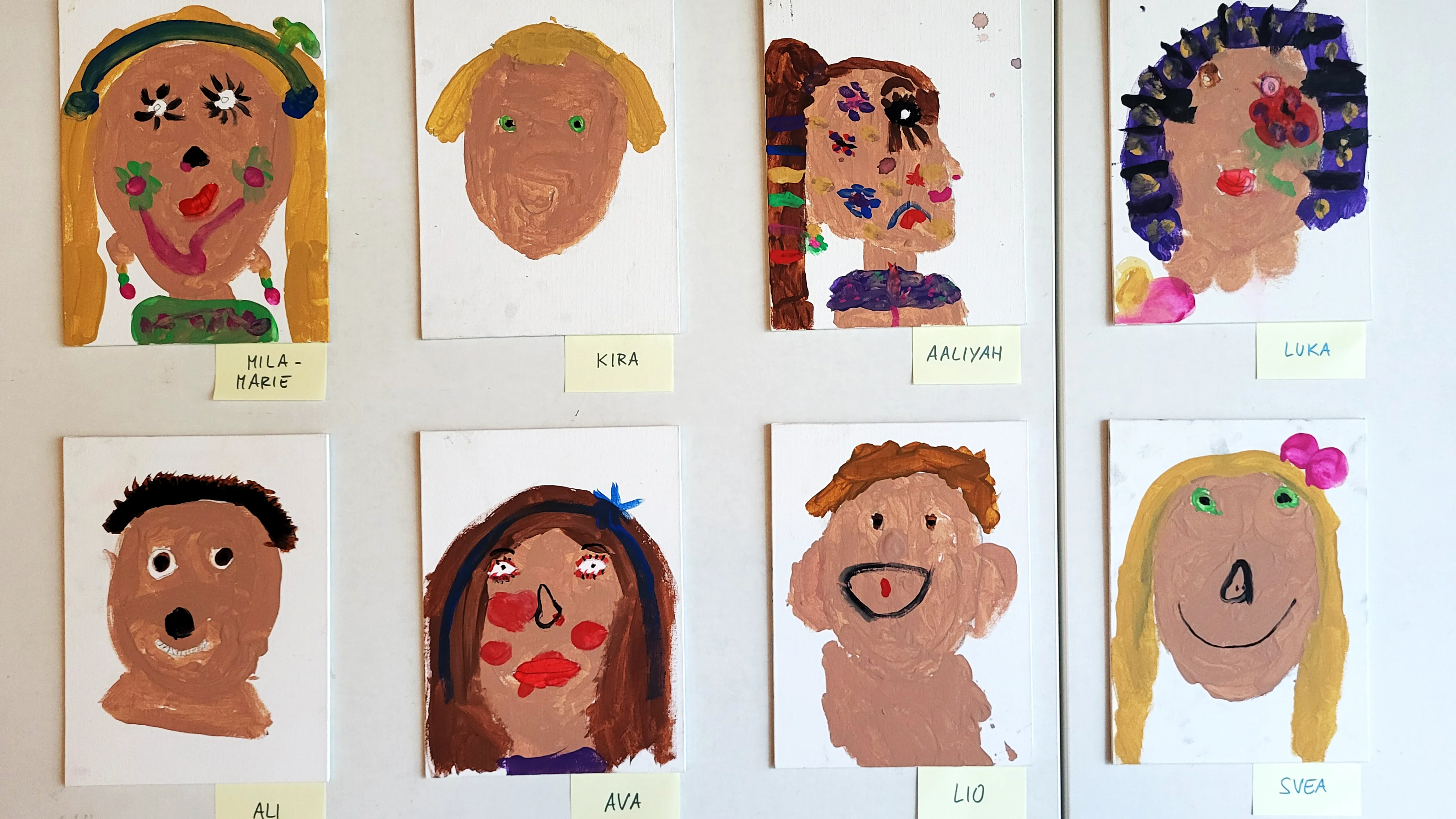 Auf dem Bild sind acht bunte Kinderporträts zu sehen, jeweils einzeln auf kleinen Leinwänden gemalt und an einer Wand aufgehängt. Unter jedem Bild klebt ein Zettel mit einem Vornamen (z.B. Mila‑Marie, Kira, Aaliyah, Luka, Ali, Ava, Lio, Svea), sodass erkennbar ist, welches Kind welches Bild gestaltet hat.