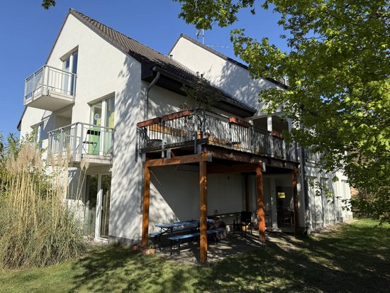 Das Bild zeigt ein zweistöckiges Haus mit einem Balkon und einer Terrasse. Das Haus hat weiße Wände und ein dunkles Dach. Es gibt mehrere Fenster und Türen, einige davon führen auf den Balkon. Die Terrasse im Erdgeschoss ist überdacht und hat Holzbalken als Stützen. Im Vordergrund sind grüne Bäume und Gras zu sehen, die teilweise Schatten auf das Haus werfen.