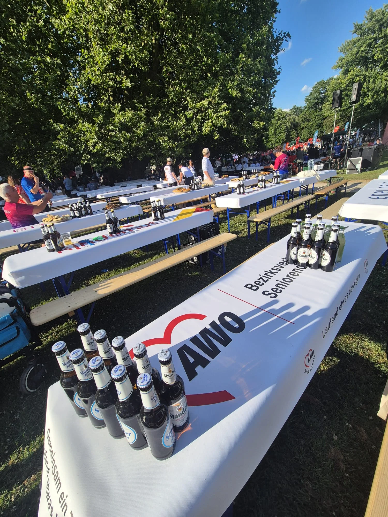 Auf dem Foto sind mehrere Biergarnituren im Freien zu sehen, die mit weißen Tischhussen und dem AWO-Logo sowie der Aufschrift des Bezirksverbands und der Seniorendienste versehen sind. Auf den vorderen Tischen stehen Reihen mit Bierflaschen, im Hintergrund sitzen und stehen Menschen unter großen Bäumen, sodass der Eindruck einer Veranstaltung oder eines Festes im Grünen entsteht.