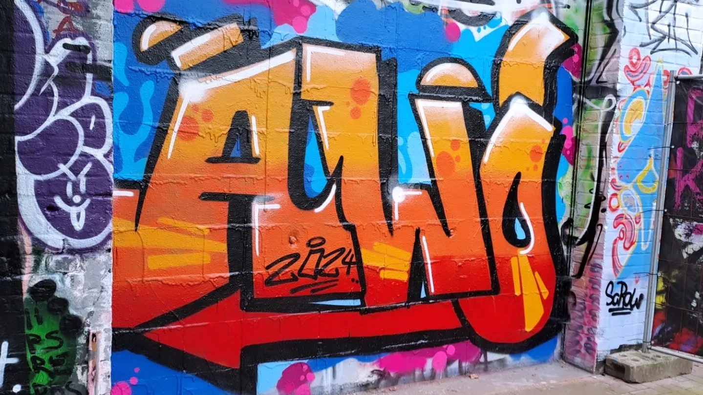 Das Foto zeigt ein Graffiti-Kunstwerk an einer Wand. Das Hauptmerkmal des Graffitis ist das Wort “AWO”, das in großen, stilisierten Buchstaben geschrieben ist. Die Buchstaben sind hauptsächlich orange mit einer weißen Umrandung und Schatteneffekten, was ihnen ein dreidimensionales Aussehen verleiht. Der Hintergrund ist mit verschiedenen anderen Graffiti-Tags und Designs in mehreren Farben gefüllt, was eine lebendige und belebte Szene schafft.