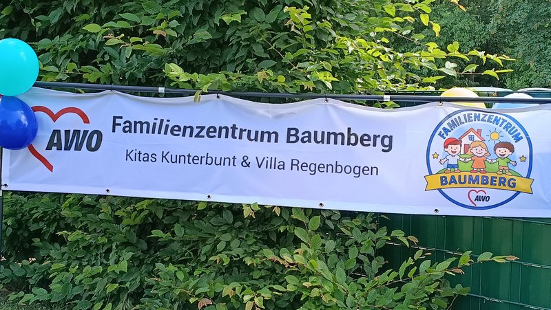 Auf dem Bild ist ein Banner des „Familienzentrums Baumberg“ der AWO zu sehen, das offenbar bei einer Außenveranstaltung aufgehängt ist. Inhalte des Banners Links befindet sich das AWO‑Logo mit dem roten Herz und dem Schriftzug „AWO“. Daneben steht in großer Schrift „Familienzentrum Baumberg“, darunter in kleinerer Zeile „Kitas Kunterbunt & Villa Regenbogen“. Rechts im Banner ist ein rundes Logo mit drei gezeichneten Kindern, Haus, Sonne und Wolken sowie dem Schriftzug „Familienzentrum Baumberg“ auf einem gelben Band. Umgebung auf dem Foto Das Banner ist an einem Metallgestell vor einer dichten grünen Hecke bzw. Buschreihe befestigt.