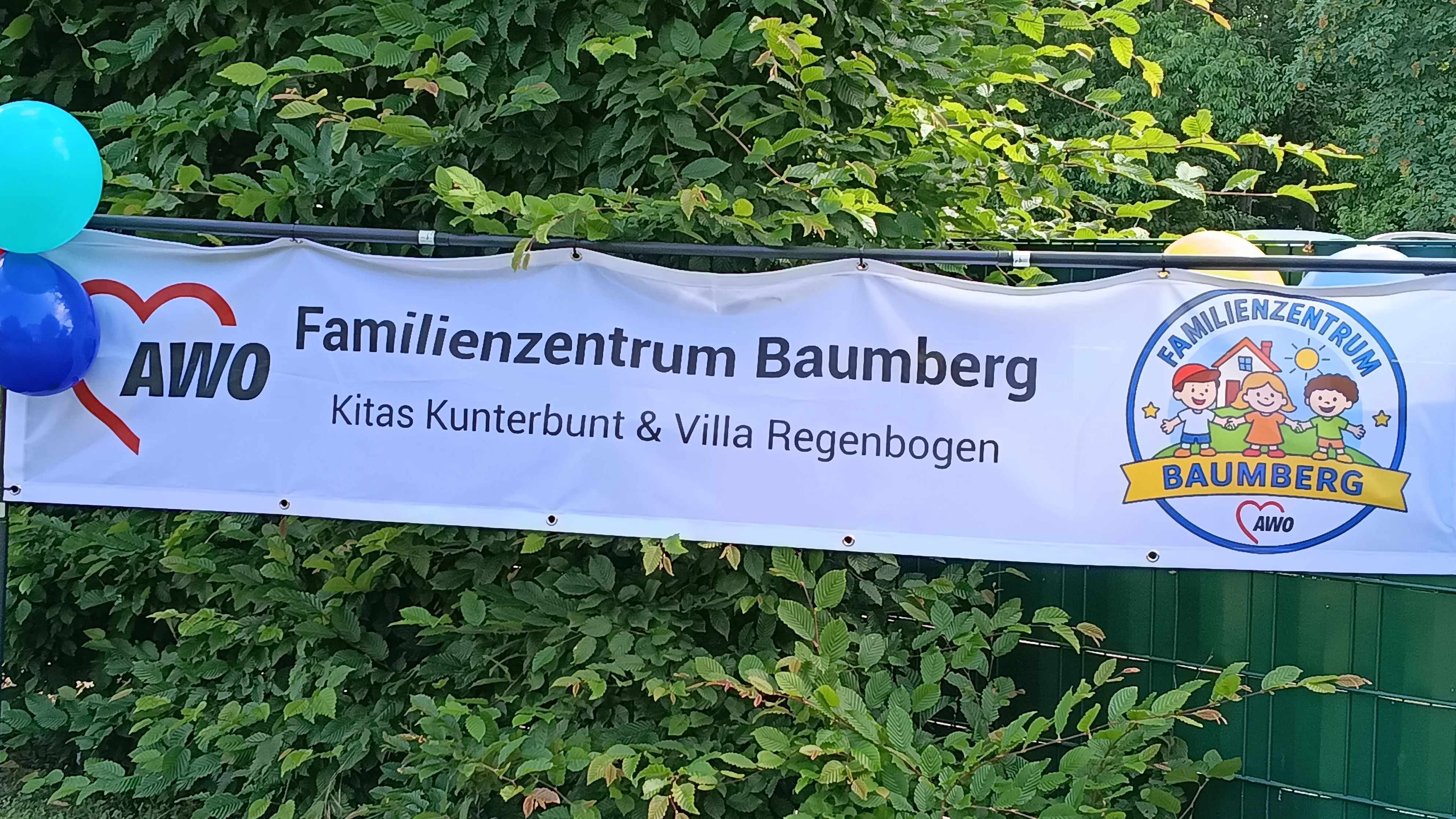 Auf dem Bild ist ein Banner des „Familienzentrums Baumberg“ der AWO zu sehen, das offenbar bei einer Außenveranstaltung aufgehängt ist. Inhalte des Banners Links befindet sich das AWO‑Logo mit dem roten Herz und dem Schriftzug „AWO“. Daneben steht in großer Schrift „Familienzentrum Baumberg“, darunter in kleinerer Zeile „Kitas Kunterbunt & Villa Regenbogen“. Rechts im Banner ist ein rundes Logo mit drei gezeichneten Kindern, Haus, Sonne und Wolken sowie dem Schriftzug „Familienzentrum Baumberg“ auf einem gelben Band. Umgebung auf dem Foto Das Banner ist an einem Metallgestell vor einer dichten grünen Hecke bzw. Buschreihe befestigt.