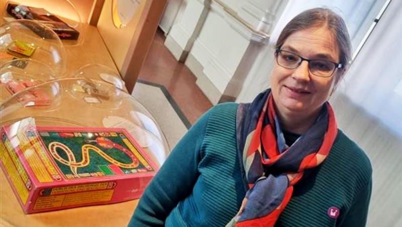 Auf dem Foto steht eine Person in einem Ausstellungsraum eines Museums oder einer ähnlichen Einrichtung. Im Hintergrund sieht man einen Holztisch mit mehreren abgedeckten Stationen und einem bunten Brettspiel mit Schlangen-Motiv unter einer transparenten Haube.