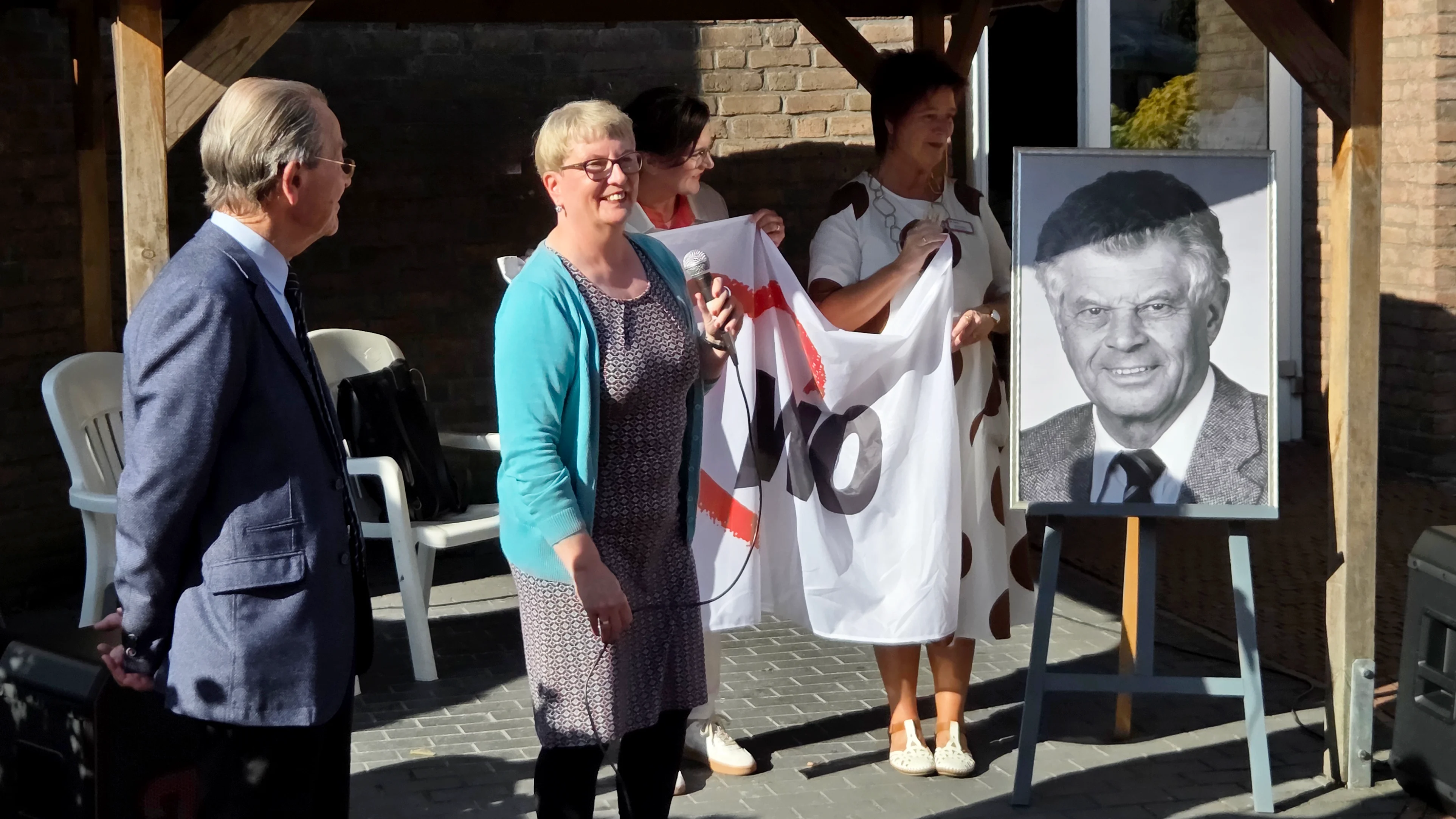 Auf dem Foto sind drei Personen zu sehen, die unter einem Unterstand stehen, vermutlich bei einer Zeremonie oder Veranstaltung im Freien. In der Mitte hält eine Frau, die eine blaue Strickjacke trägt, eine Art Fahne oder Banner, auf dem die Buchstaben "AWO" zu erkennen sind. Neben ihr, auf ihrer rechten Seite, steht eine andere Frau, die ein weißes, gepunktetes Kleid trägt und ebenfalls das Banner festhält. Eine dritte Person, ein Mann in einem blauen Anzug, steht auf der linken Seite. Neben den Personen steht eine Staffelei mit einem gerahmten Porträtfoto eines Mannes in einem Anzug. Die Szene spielt sich vor einer Ziegelsteinwand ab, und im Hintergrund sind weiße Plastikstühle zu sehen.