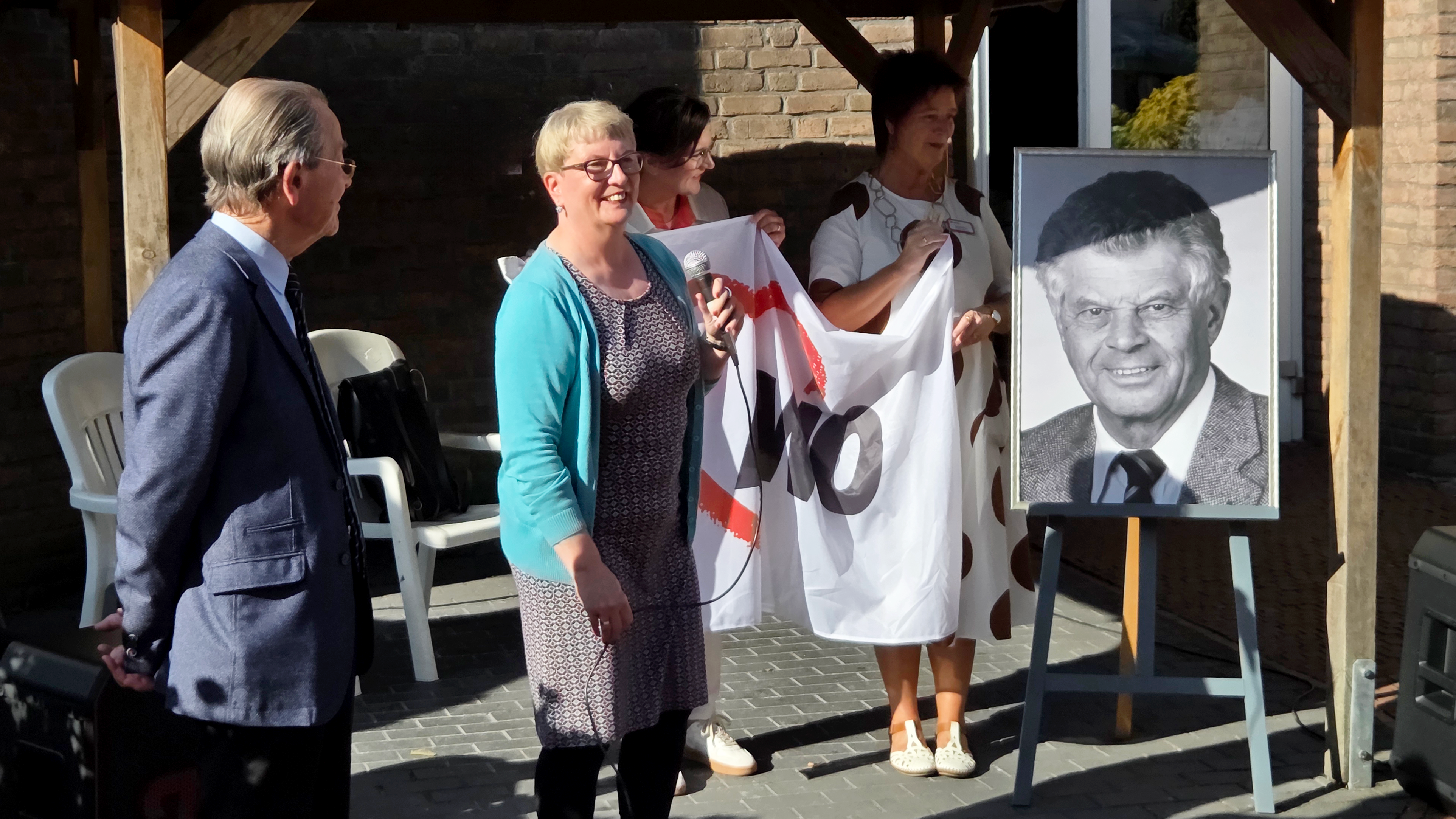 Auf dem Foto sind drei Personen zu sehen, die unter einem Unterstand stehen, vermutlich bei einer Zeremonie oder Veranstaltung im Freien. In der Mitte hält eine Frau, die eine blaue Strickjacke trägt, eine Art Fahne oder Banner, auf dem die Buchstaben "AWO" zu erkennen sind. Neben ihr, auf ihrer rechten Seite, steht eine andere Frau, die ein weißes, gepunktetes Kleid trägt und ebenfalls das Banner festhält. Eine dritte Person, ein Mann in einem blauen Anzug, steht auf der linken Seite. Neben den Personen steht eine Staffelei mit einem gerahmten Porträtfoto eines Mannes in einem Anzug. Die Szene spielt sich vor einer Ziegelsteinwand ab, und im Hintergrund sind weiße Plastikstühle zu sehen.