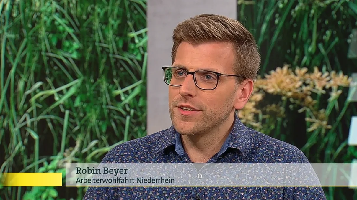 Die Person trägt ein blaues Hemd mit einem Muster und befindet sich in einem Interview. Im unteren Drittel des Bildes steht der Text “Robin Beyer Arbeitswohlfahrt Niederrhein”, was darauf hindeutet, dass diese Person Robin Beyer heißt und mit einer Organisation namens Arbeitswohlfahrt Niederrhein in Verbindung steht.