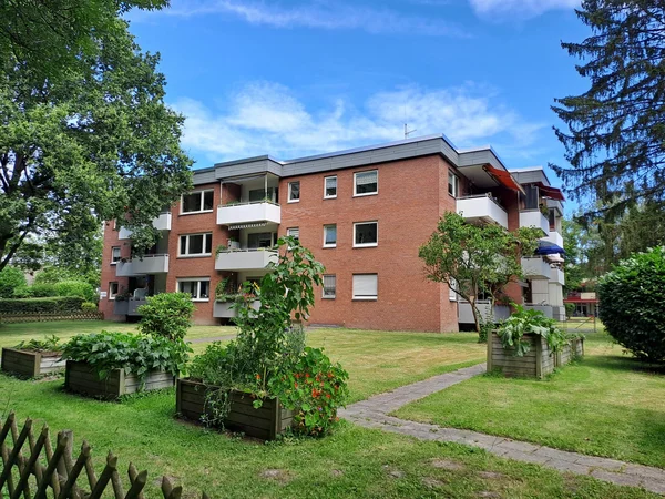 Auf dem Bild ist ein mehrstöckiges Wohnhaus aus rotem Klinker mit mehreren Balkonen zu sehen, das in einer grünen Gartenanlage steht. Im Vordergrund erkennt man Hochbeete mit verschiedenen Pflanzen sowie eine Rasenfläche mit einem gepflasterten Weg, umgeben von Bäumen und Büschen.