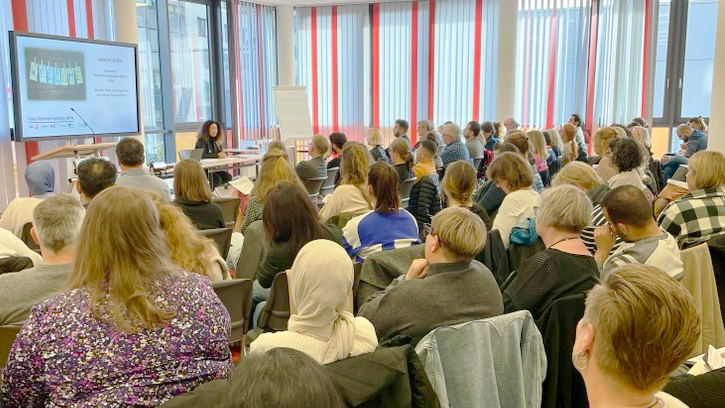 Auf dem Foto ist eine größere Gruppe von Menschen zu sehen, die in einem Seminar- oder Konferenzraum an einer Präsentation teilnehmen. Die Zuhörer sind in Reihen angeordnet und blicken auf eine Rednerin oder einen Redner, der oder die vorne an einem Tisch sitzt und eine Präsentation hält, die auf einem großen Bildschirm gezeigt wird. Die Präsentation scheint sich auf ein Projekt oder eine Veranstaltung zu beziehen, und der Raum ist modern gestaltet, mit großen Fenstern im Hintergrund, die mit rot-weiß gestreiften Vorhängen versehen sind.