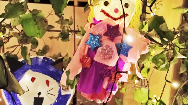 Auf dem Foto sind zwei handgefertigte Figuren zu sehen, die anscheinend zur Dekoration aufgehängt wurden, vermutlich für ein Fest wie das Laternenfest, wie der Dateiname andeutet. Hier sind die Details: Puppe/Figur: Im Zentrum hängt eine Puppen-ähnliche Figur mit gelbem "Haar", einem lächelnden Gesicht (roter Mund, schwarze Augen, rote Wangen) und einem Körper aus buntem (rosa/lila) Material, verziert mit glitzernden Sternen. Löwen-Maske: Links davon ist eine Figur in Form eines Löwenkopfs zu sehen, die auf einen blauen und weißen Pappteller gemalt oder geklebt wurde. Sie hat ein aufgerissenes Maul und rote Details. Lichterkette und Blätter: Die Figuren sind in einem Gewirr aus grünen Blättern und einer Lichterkette mit kleinen, leuchtenden Punkten angebracht, was eine warme, festliche Atmosphäre schafft.