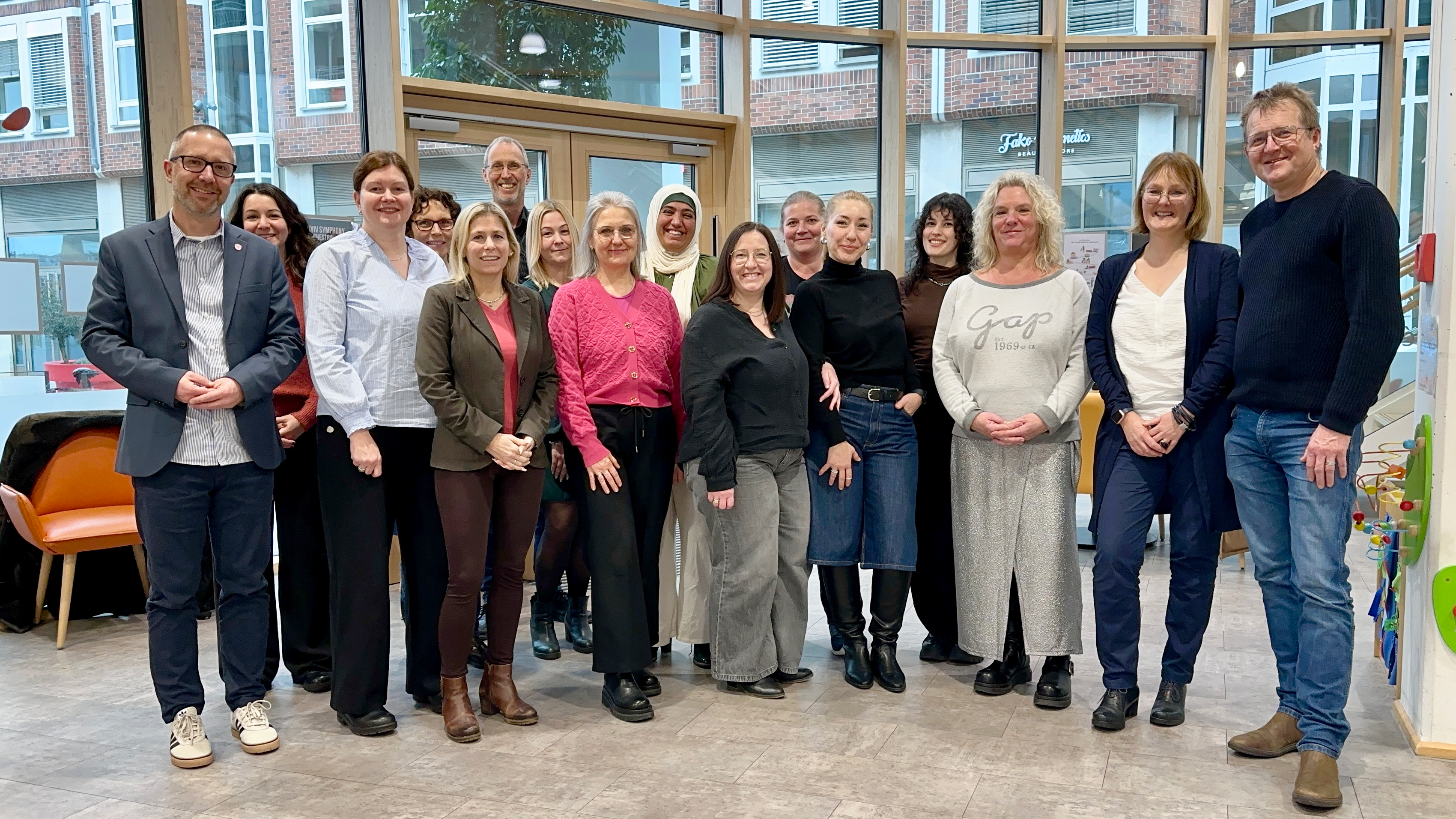 Auf dem Foto steht eine größere Gruppe von Erwachsenen in einem hellen Raum mit großen Fenstern, vermutlich ein Team oder eine Arbeitsgruppe. Die Personen posieren in einer Reihe für ein Gruppenbild; im Hintergrund sind Stadtgebäude und etwas Einrichtung wie Stühle und Spielzeug zu sehen.