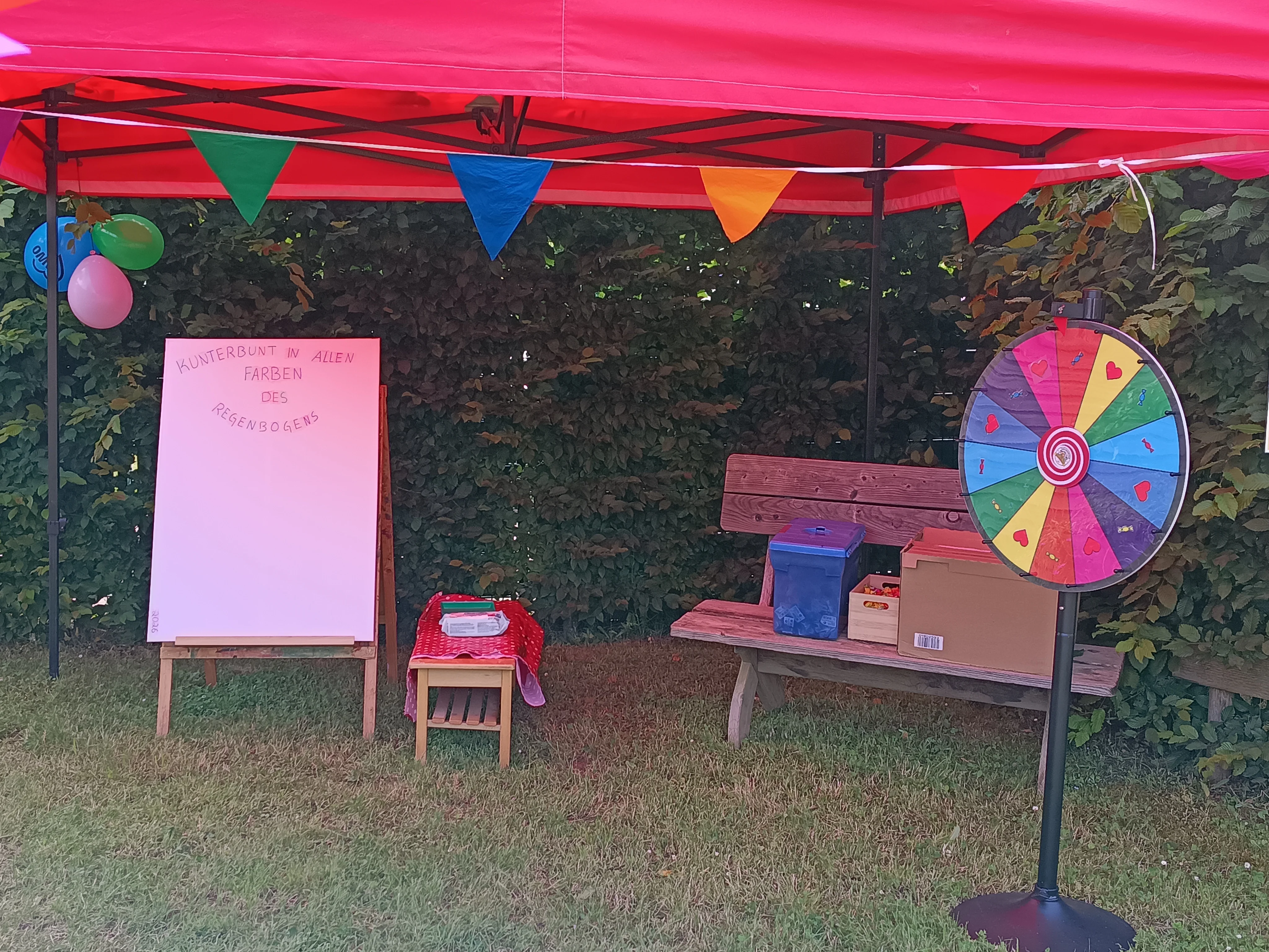 Auf dem Bild ist ein kunterbunt gestalteter Aktionsstand auf einer Wiese unter einem roten Pavillon zu sehen. Elemente des Standes Links steht eine große weiße Tafel auf einem Holzständer, oben mit dem Schriftzug „Kunterbunt in allen Farben des Regenbogens“. Daneben steht ein kleiner Tisch mit rot‑weiß karierter Decke und Materialien darauf. Umgebung und Deko Rechts im Bild befindet sich ein bunt bemaltes Glücksrad mit Herzmotiven vor einer Holzbank, auf der Kisten und Boxen stehen. Unter dem Pavillon hängen bunte Wimpelketten und Ballons, im Hintergrund ist eine dichte grüne Hecke zu erkennen