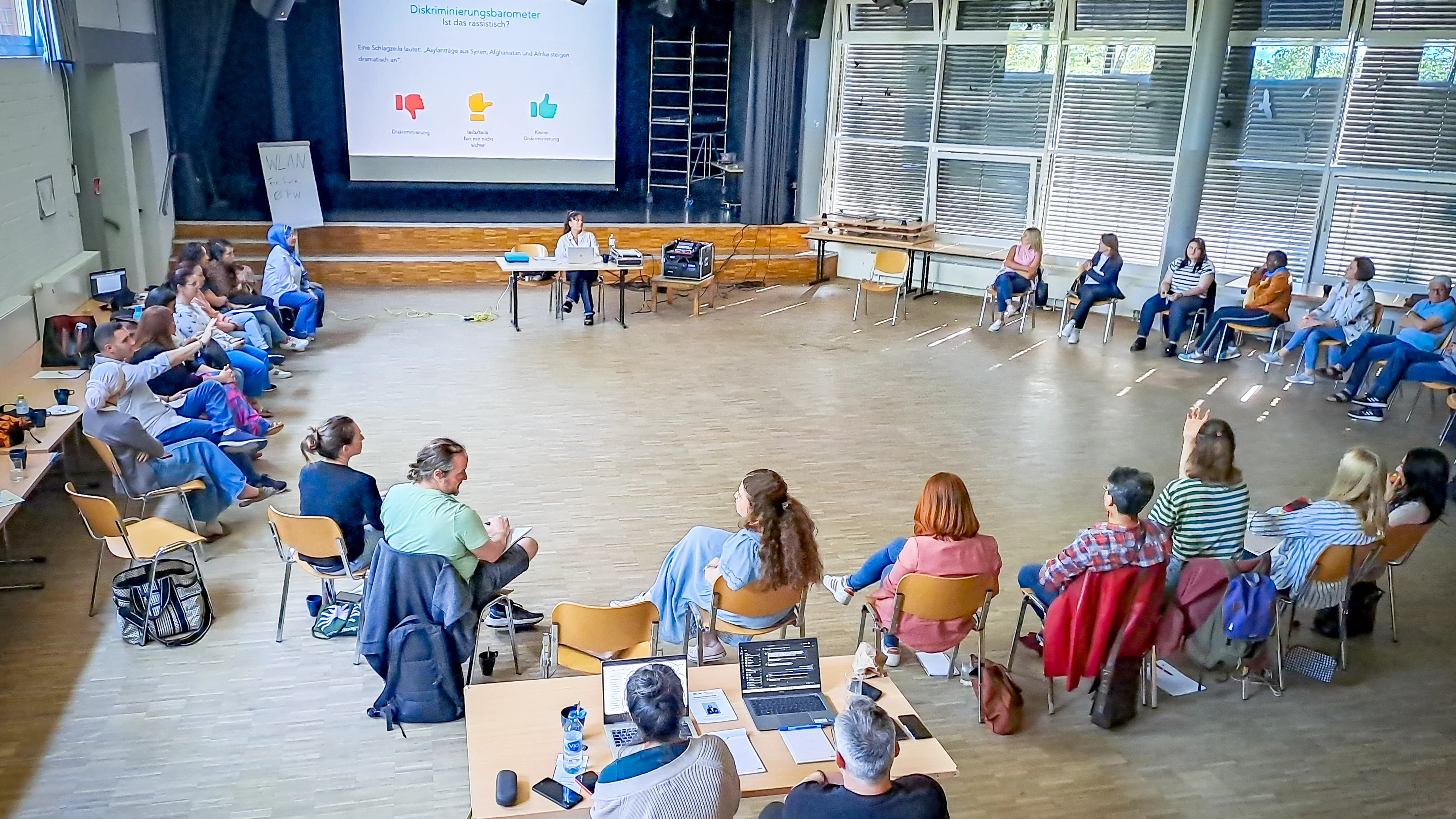 Auf dem Foto ist eine Gruppe von Menschen zu sehen, die in einem großen, kreisförmig angeordneten Raum an einer Sitzung oder einem Workshop teilnehmen. Sie sitzen auf Stühlen, die in einem Kreis aufgestellt sind. In der Mitte des Raumes befinden sich zwei Tische mit Laptops und anderen Materialien. An der Vorderseite des Raumes steht ein Bildschirm mit einer Präsentation, die den Titel „Diskriminierungsbarometer“ trägt und die Frage stellt: „Ist das rassistisch?“ Es gibt drei Reaktionsmöglichkeiten auf dem Bildschirm: „Daumen runter“, „Daumen zur Seite“ und „Daumen hoch“.