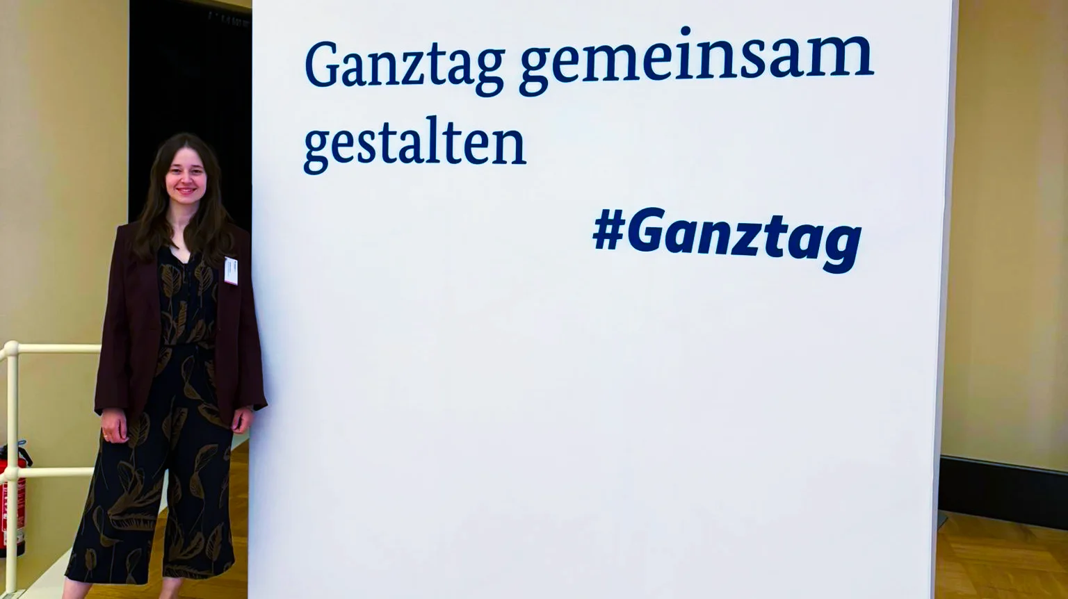 Auf dem Bild sieht man eine große weiße Stellwand mit einem Schriftzug zum Thema Ganztagsbildung sowie eine Person, die links daneben steht. Text auf der Stellwand Auf der Stellwand steht oben links das Logo und der Schriftzug des „Bundesministeriums für Bildung, Familie, Senioren, Frauen und Jugend“. In der Mitte ist der Slogan „Ganztag gemeinsam gestalten“ groß zu lesen, darunter der Hashtag „#Ganztag“.