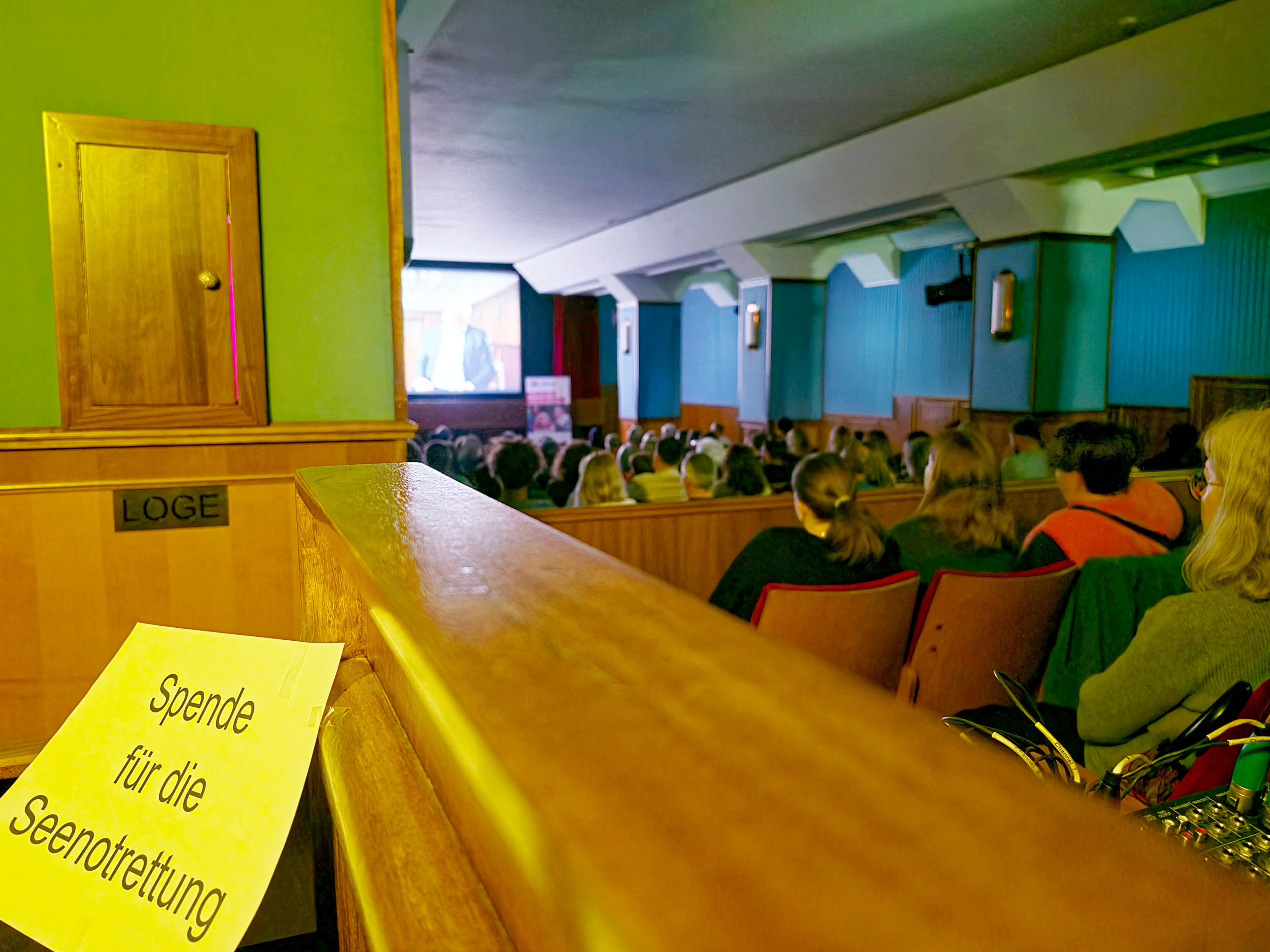 Auf dem Bild ist ein kleiner Saal bzw. ein Kinoraum mit vielen sitzenden Personen zu sehen, die auf eine Leinwand vorne im Raum blicken. Links im Vordergrund ist eine hölzerne Brüstung mit dem Schild „Spende für die Seenotrettung“ zu erkennen, außerdem ein „LOGE“-Schild an der Wand. In der Bildmitte und im Hintergrund sitzen zahlreiche Zuschauer in Reihen, der Raum hat türkisfarbene Wandverkleidungen und Holzvertäfelung. Die Leinwand vorne zeigt eine unscharf erkennbare Projektion.
